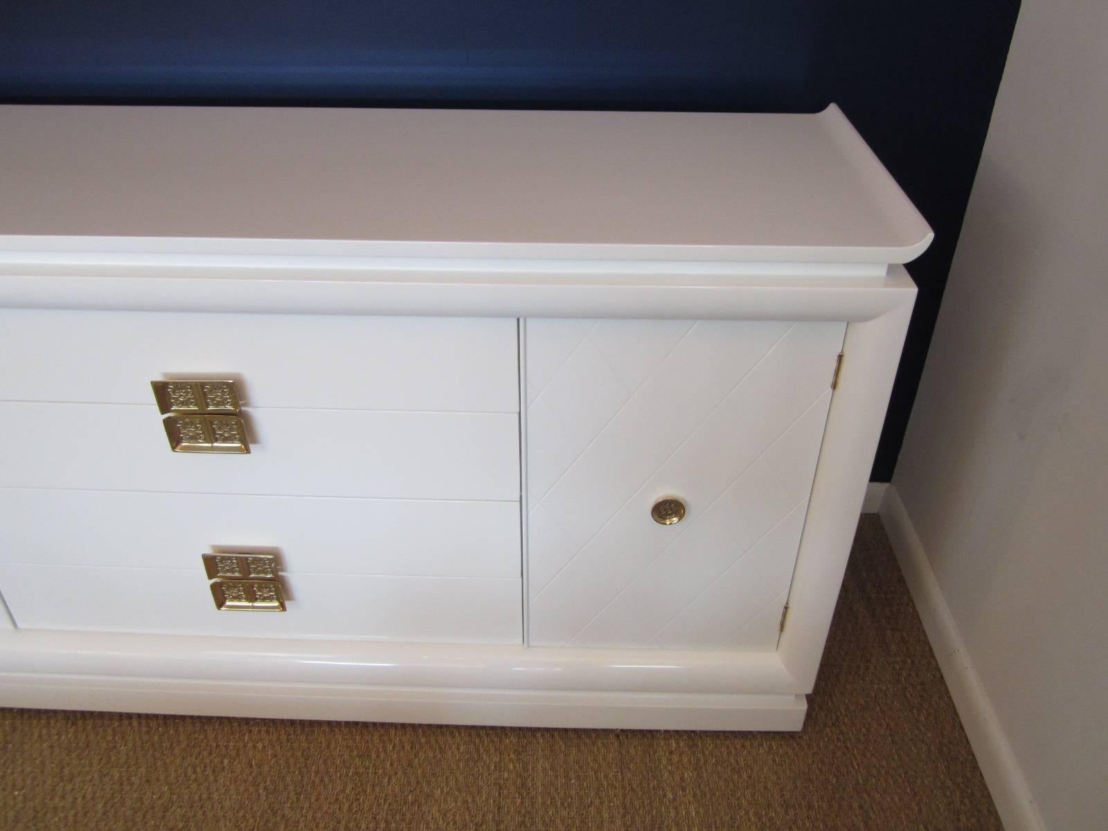 Gorgeous high gloss white lacquered credenza with original polished brass incised hardware on doors and drawers. The doors reveal bottom shelf and adjustable center shelf storage cubbies. Credenza has great 