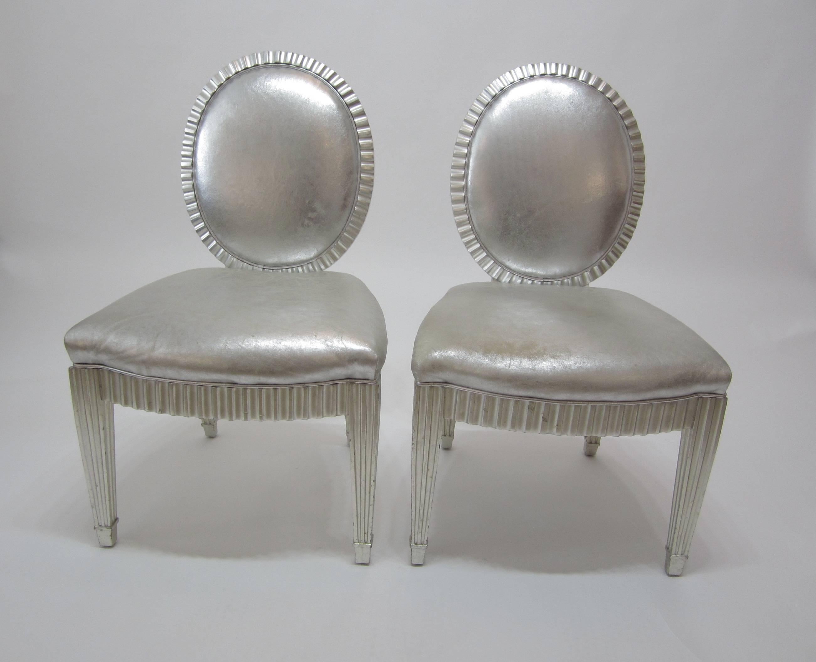 Designed by John Hutton for Donghia, these neoclassical style wood-frame side chairs are silver leafed and have been covered in silver leather metallic. Even after use with minimal but visible wear, these chairs stand out due to their unique yet