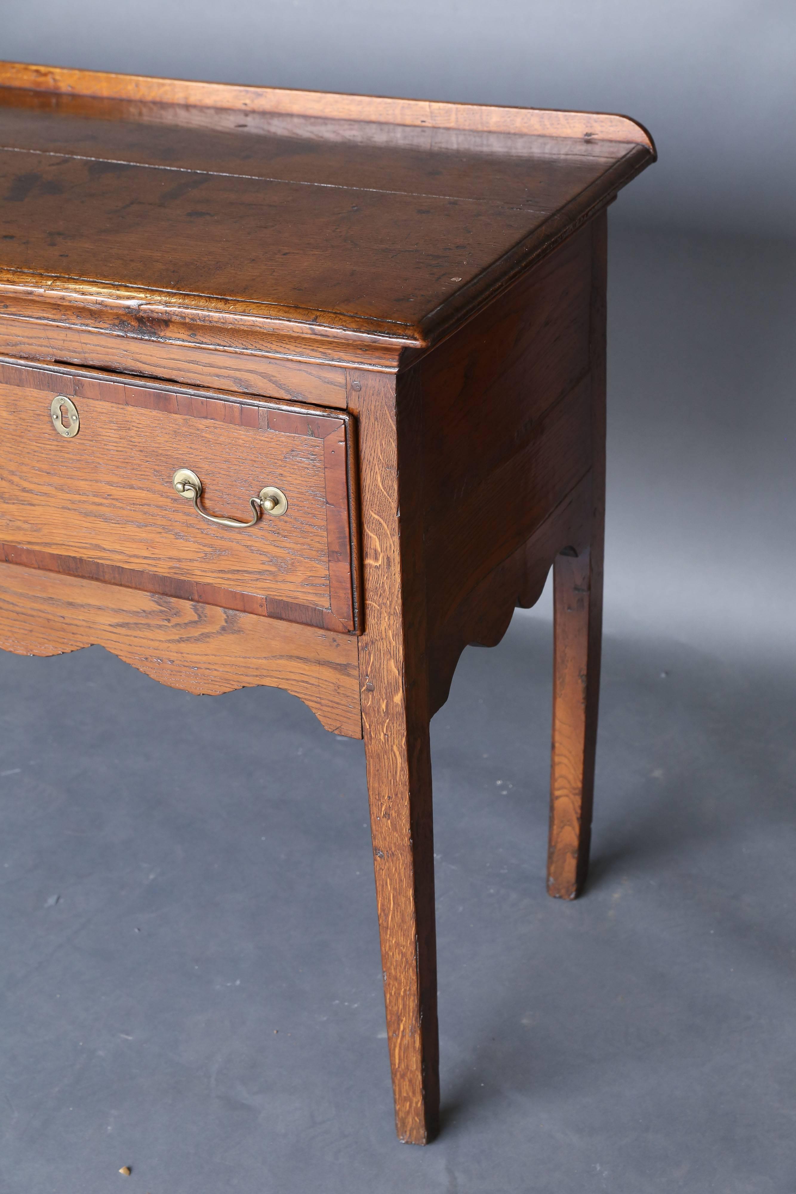 A George III oak and mahogany banded dresser base, fitted with three deep drawers, shaped apron, square tapering legs and swan neck brass pulls.