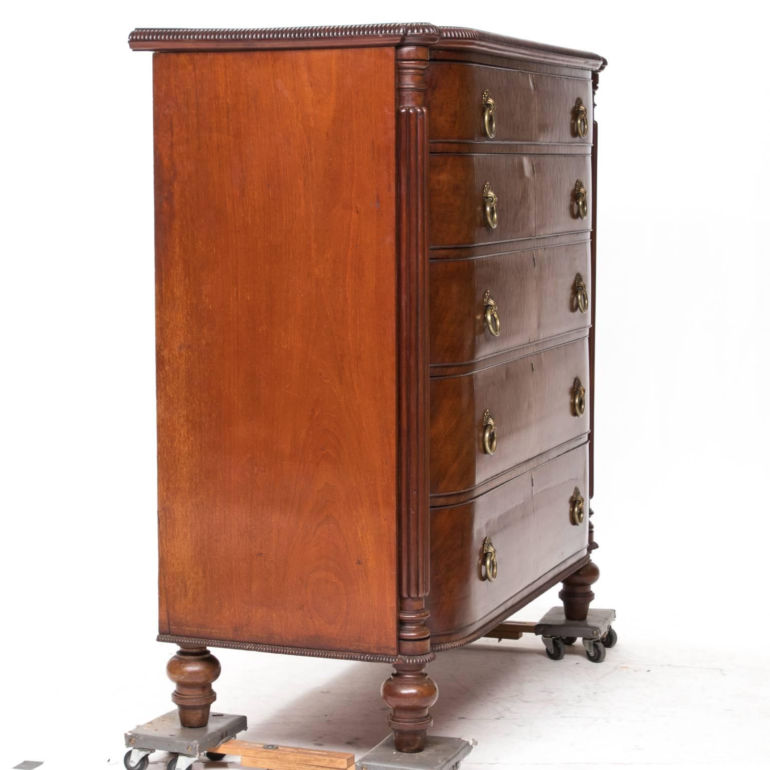 19th C. English mahogany chest of drawers in the Regency style. The front has a bow front and turned fluted columns corners. Notice the moldings present on the top and base are very defined. There are five graduated drawers with substantial brass