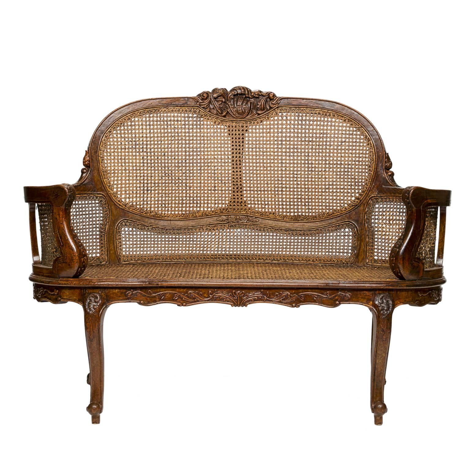 This Louis XV walnut settee features an exposed cane and carved wood frame. It has a floral cresting and carved apron. The back barrels into the arm supports, which are carved. The settee rests upon four cabriole legs. The front left panel behind