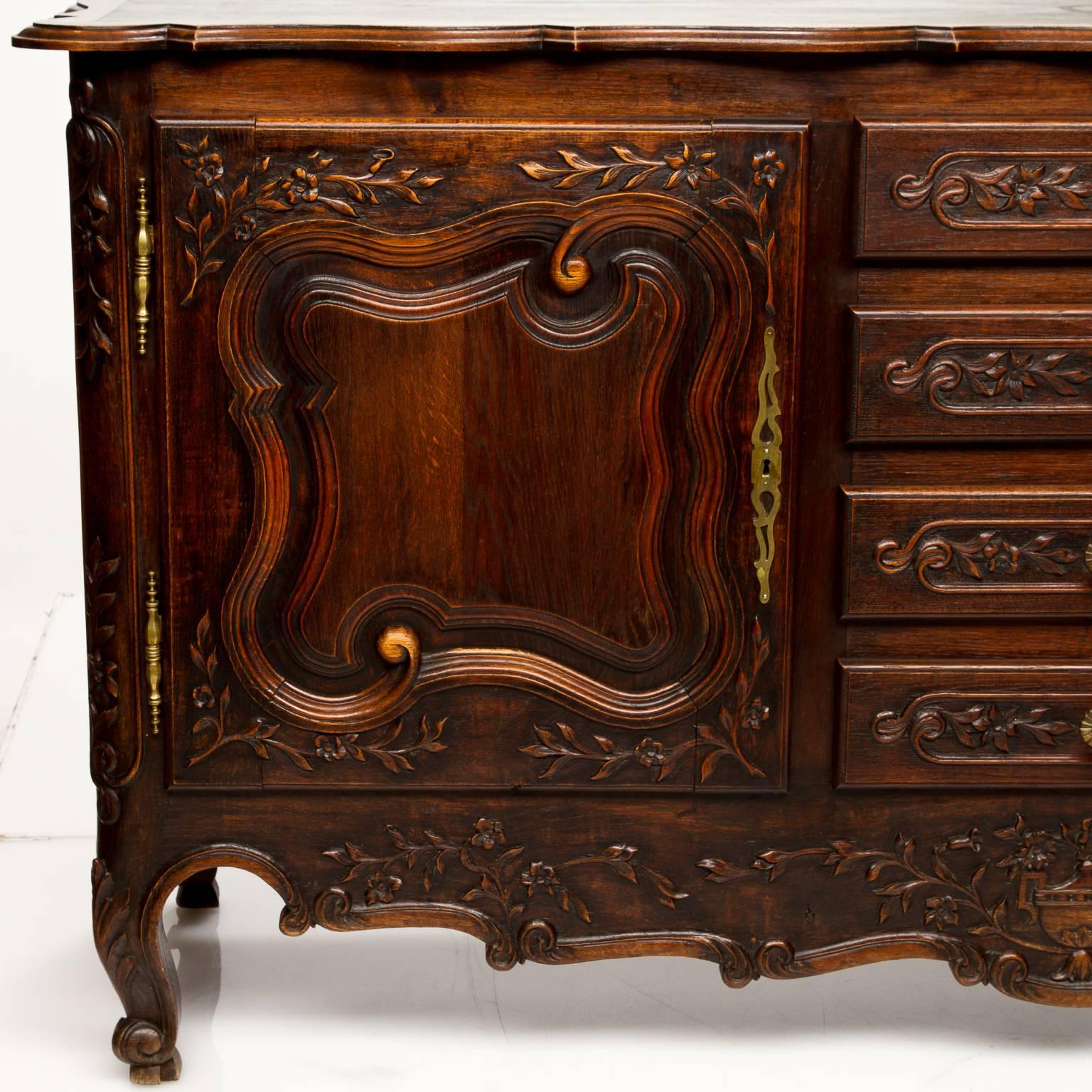 A French Provincial oak and elm 19th century buffet. This buffet has elaborate carvings, a leaf and vine with florals, shaped top, large scroll raised panel doors divided by a bank of four drawers, the apron is centered with a pastoral floral basket