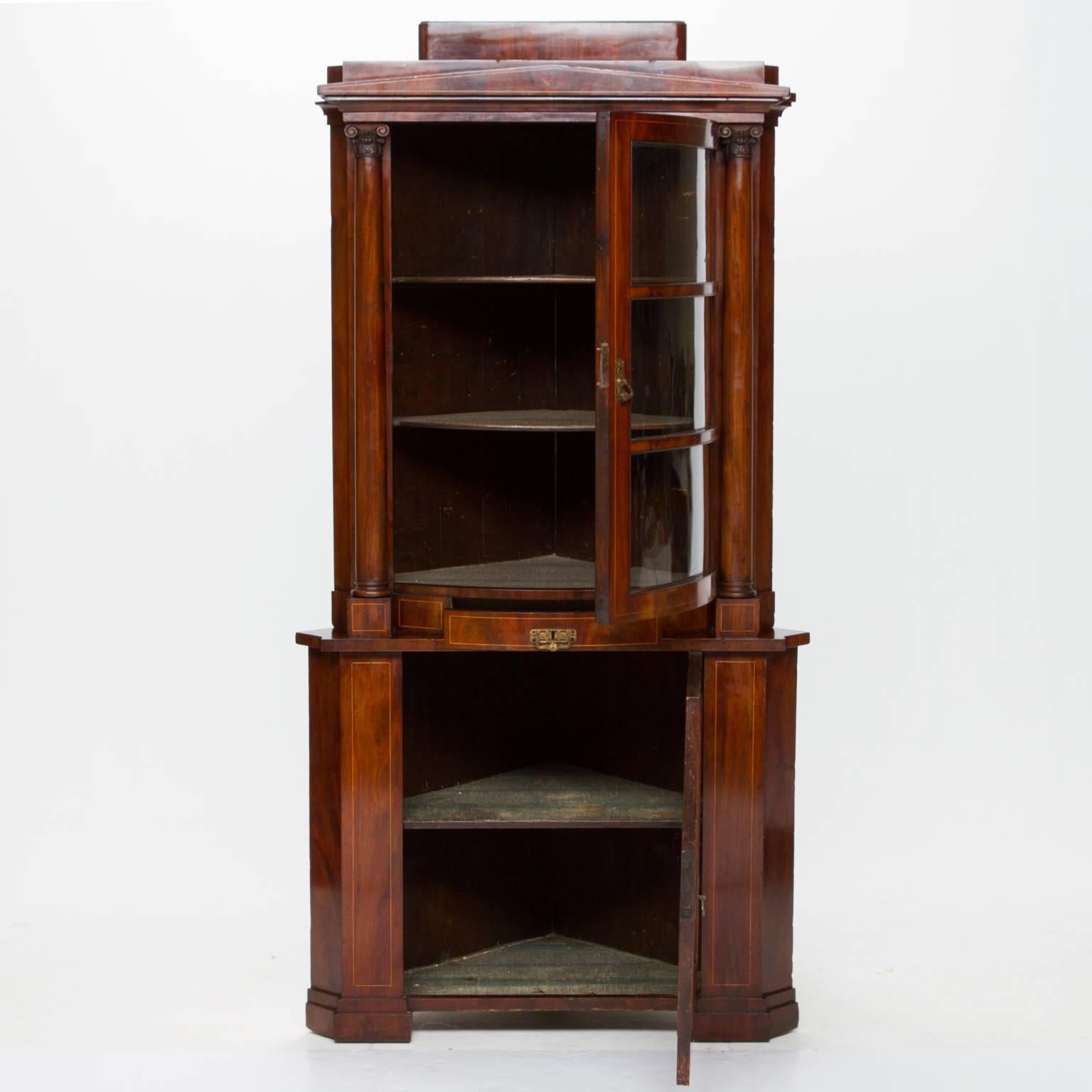 19th century mahogany inlaid English corner cabinet. This corner cabinet is two parts. The top section has a bow front. The glass is bowed and wavy. This opens to reveal two shelves with vennered fronts. On each side are two fine turned columns