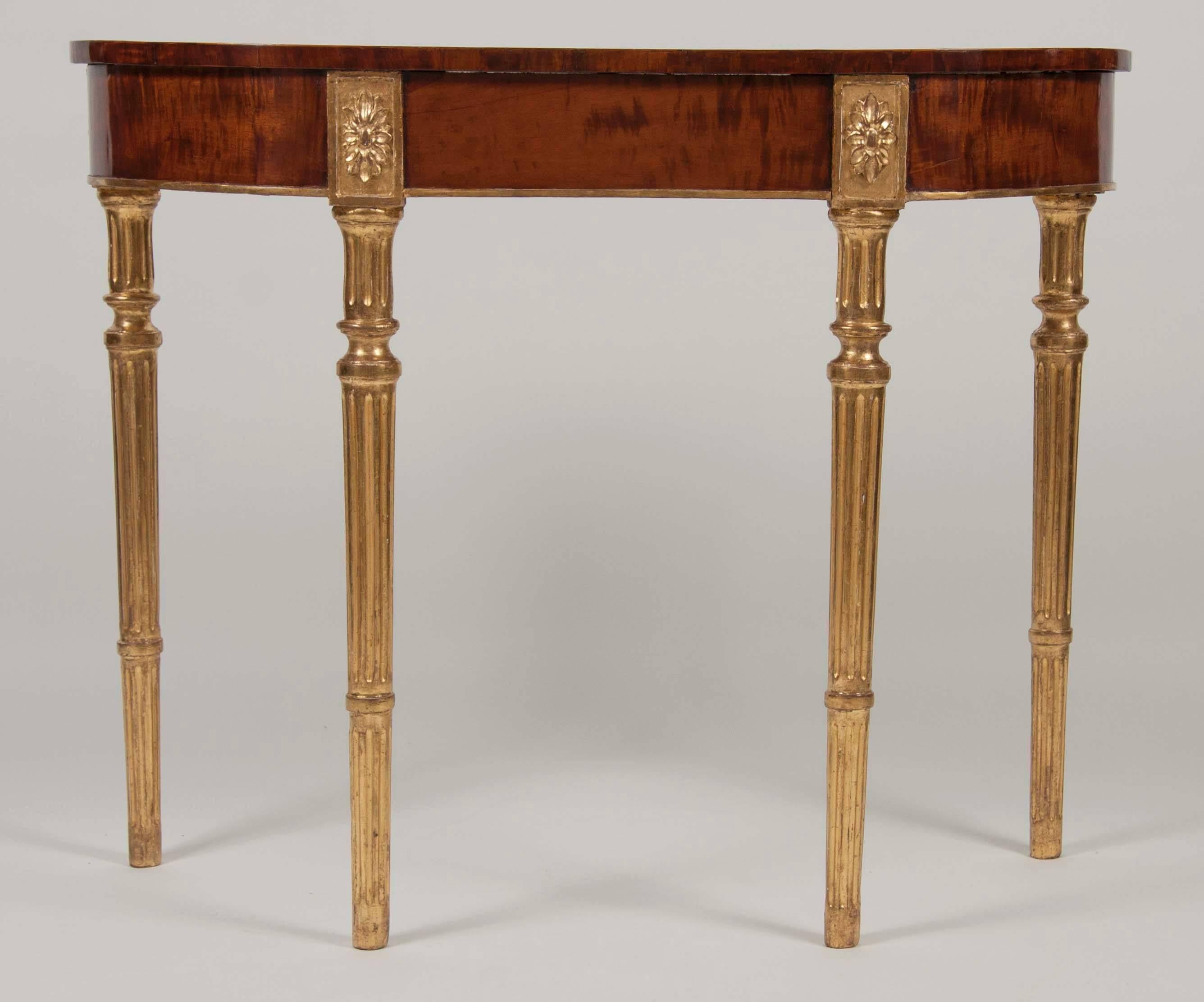 A stunning pair of matched George III period, mahogany and satinwood, parcel-gilt console tables having crossbanded 'D' form tops over gilded tapering, ring turned fluted legs.