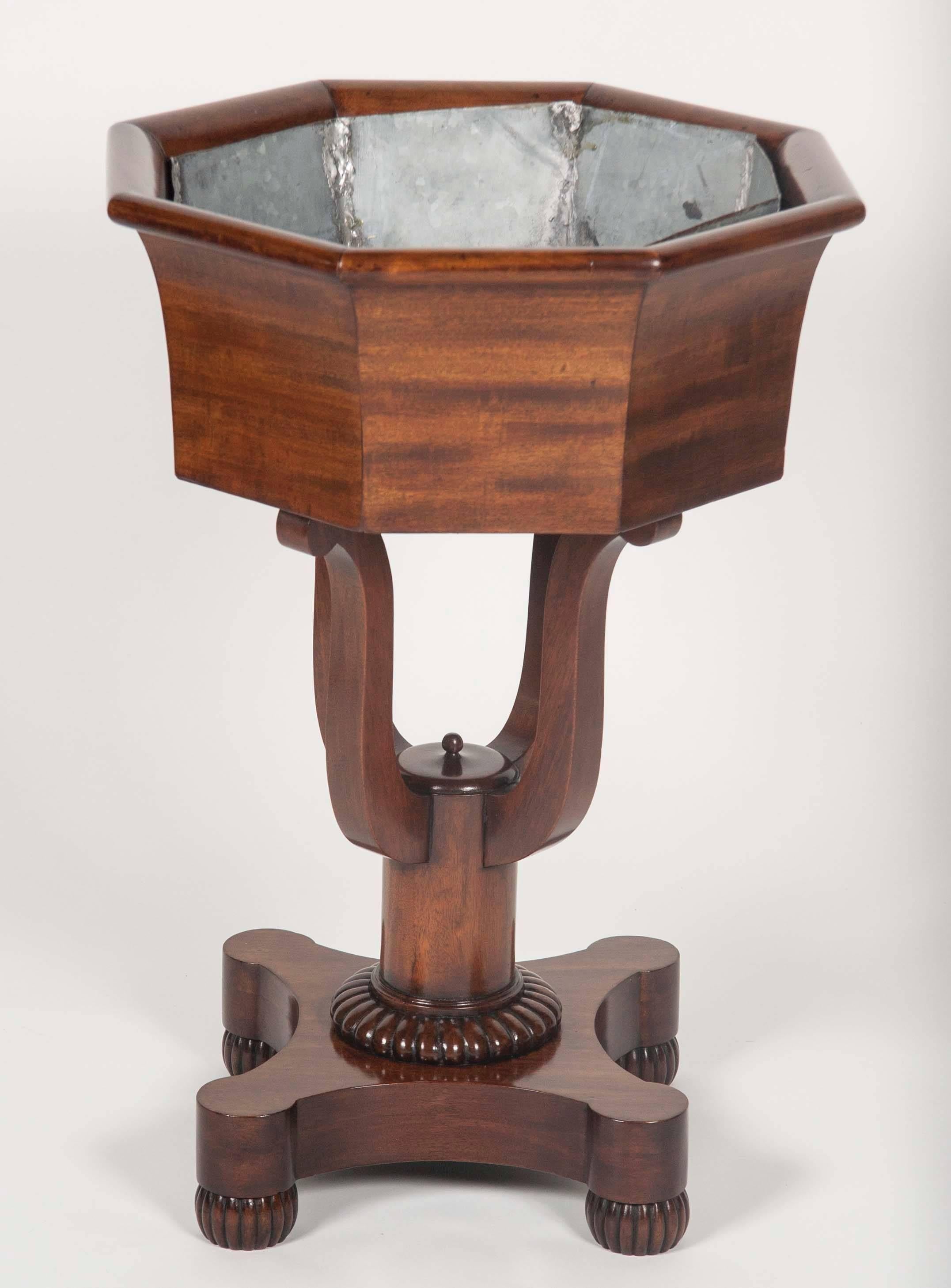 An American mahogany octagonal faceted jardinière with galvanized liner. The planter supported by four scroll form arms jointed to a single cylindrical plinth base terminating in a gadrooned pedestal on four gadrooned bun feet.