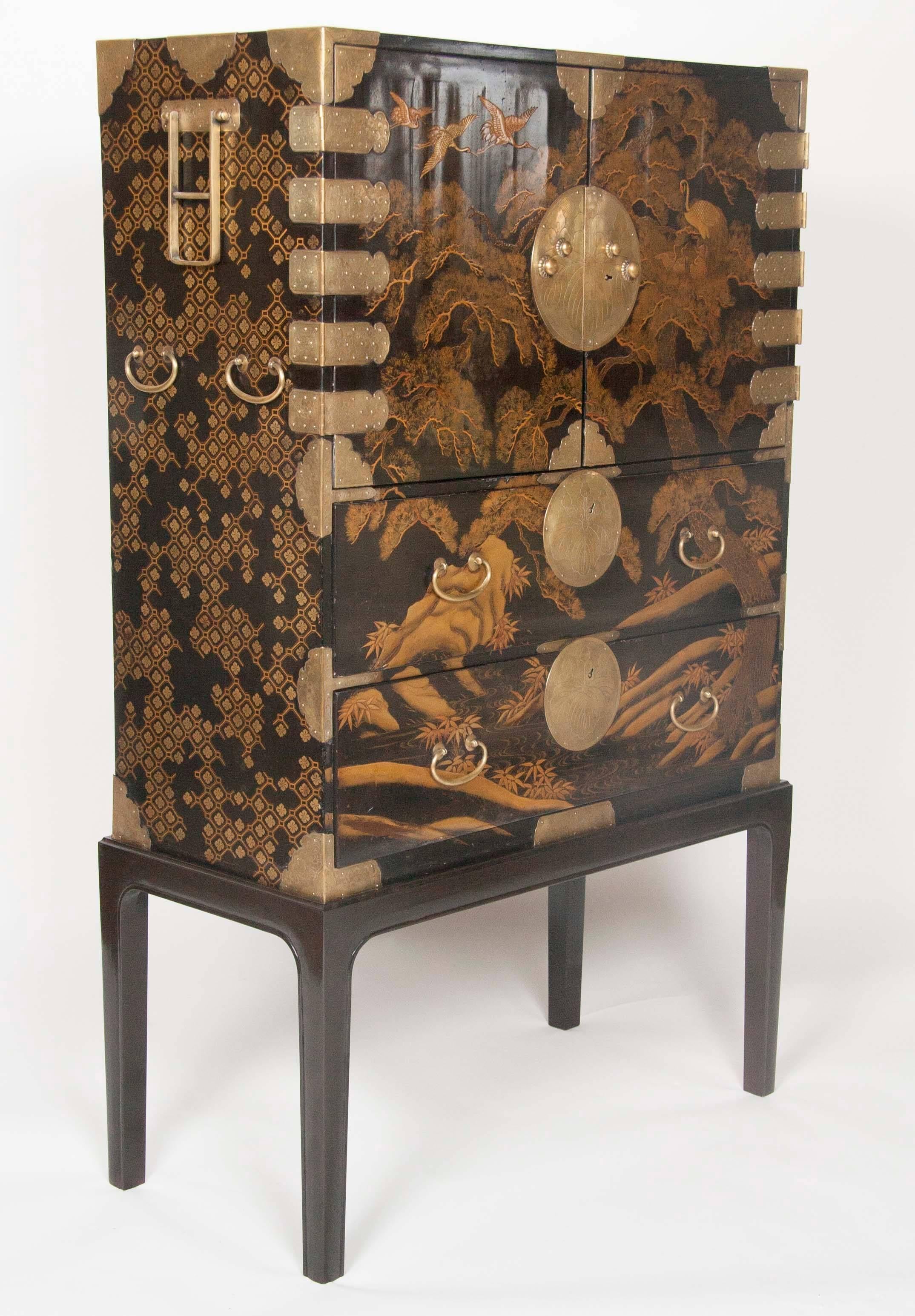 A striking early 19th century Japanese gilt and black lacquer cabinet with brass mountings; two upper doors; revealing three highly decorated lacquered Kimono trays over three ebonized Kimono trays. Two lower drawers showing cranes in pine trees.