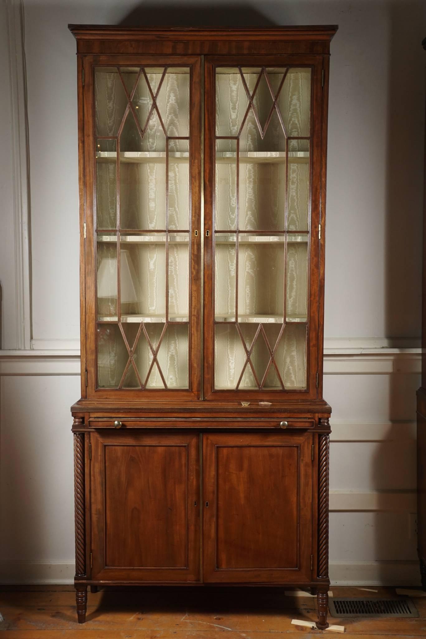 A pair of English mahogany bookcases. Moulded cornice over an upper section with two glazed doors with fabric lined back and adjustable shelves. Brushing slide with velvet surface over a lower section with two cabinet doors with shelves. Carved