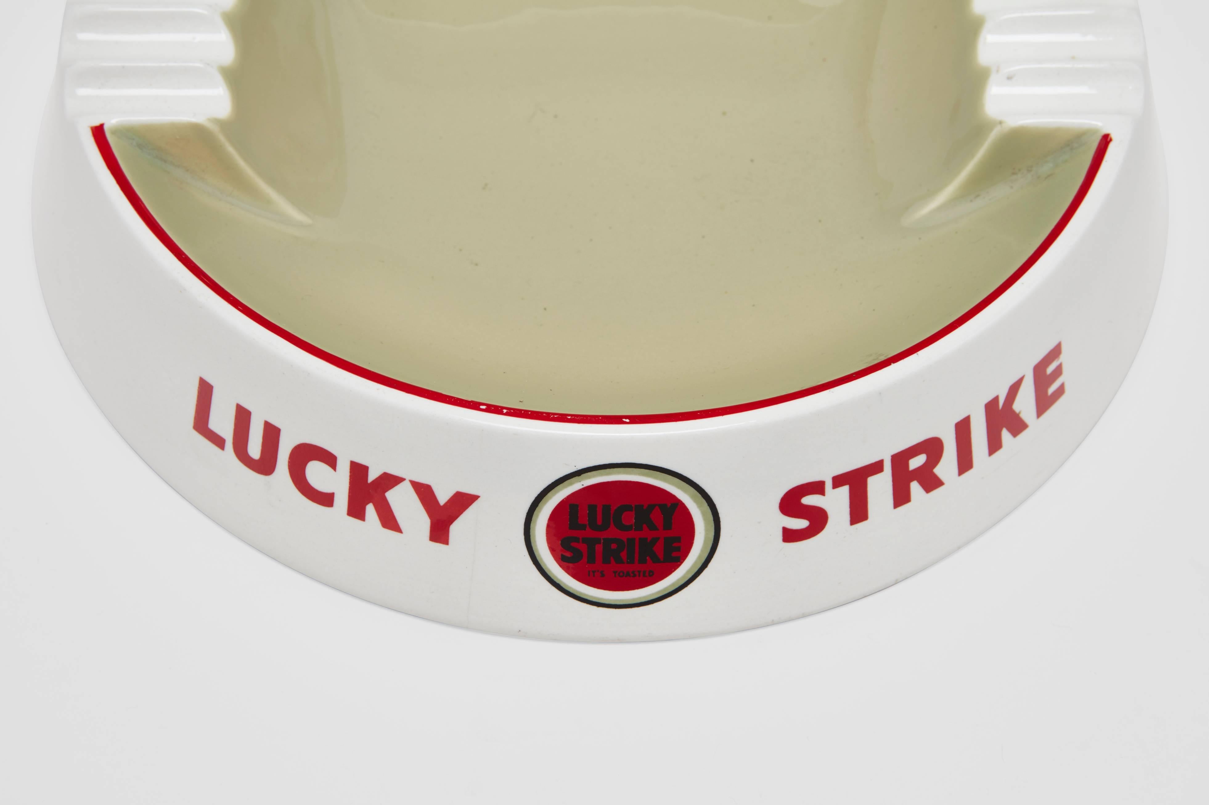 A large porcelain cigar or cigarette ashtray. This wonderful and unusually large-scale piece is a very graphic piece of Mid-Century advertising. The white border is marked either side with the 'Lucky Strike' cigarette company branding and centered
