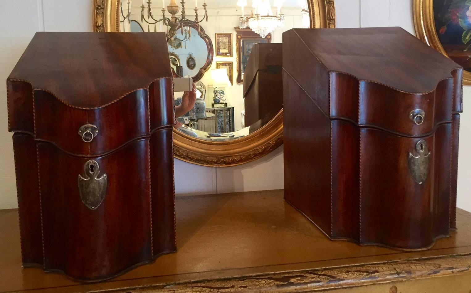 Two handsome matching George III mahogany cutlery boxes of typical form with a sloping lid, particularly bold serpentine front and original brass escutcheon and ring-handle.  The interiors do not have felt dividers to organize silverware, but are