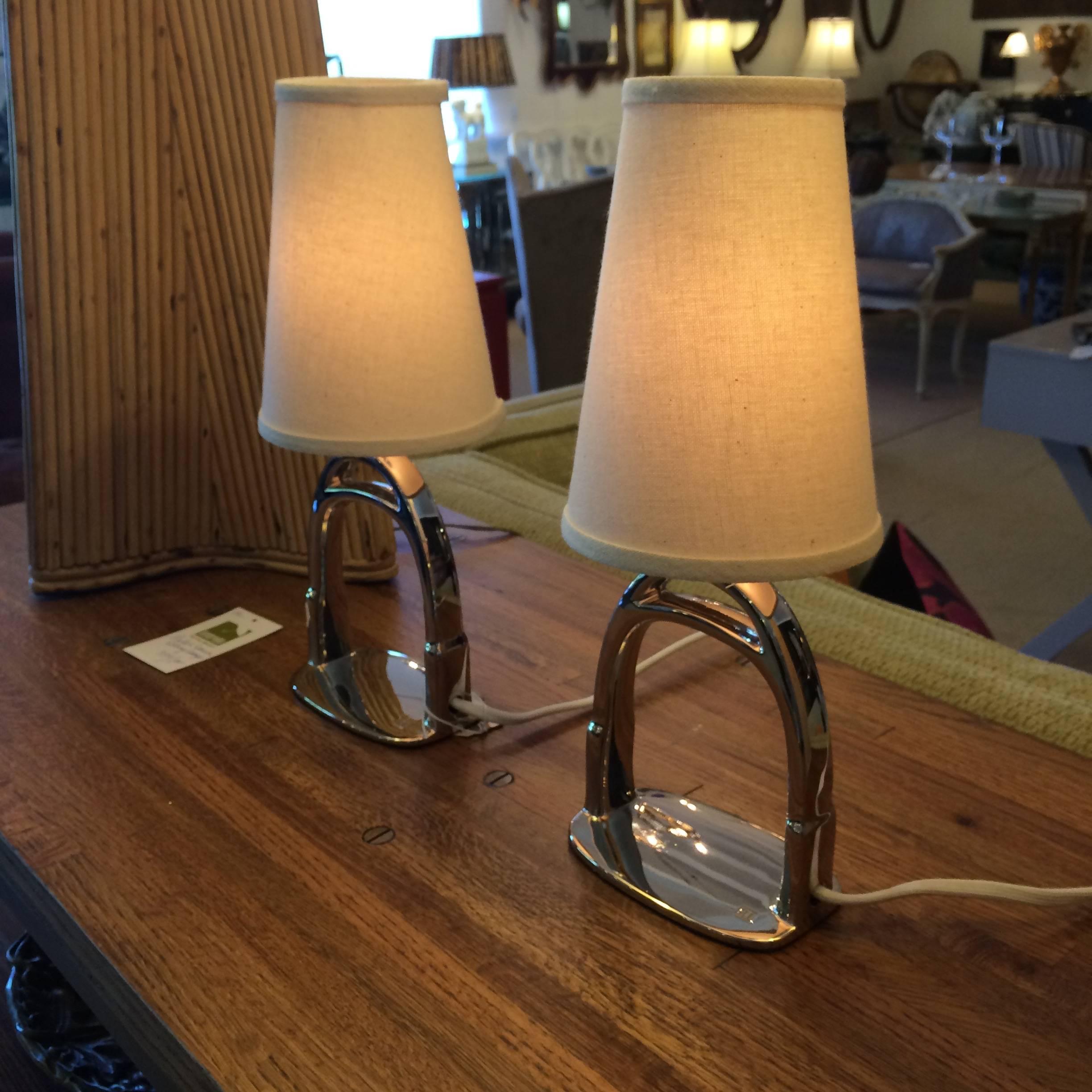 Fun and glamorous pair of small lamps with bases in the shape of chrome stirrups with off white shades. 
