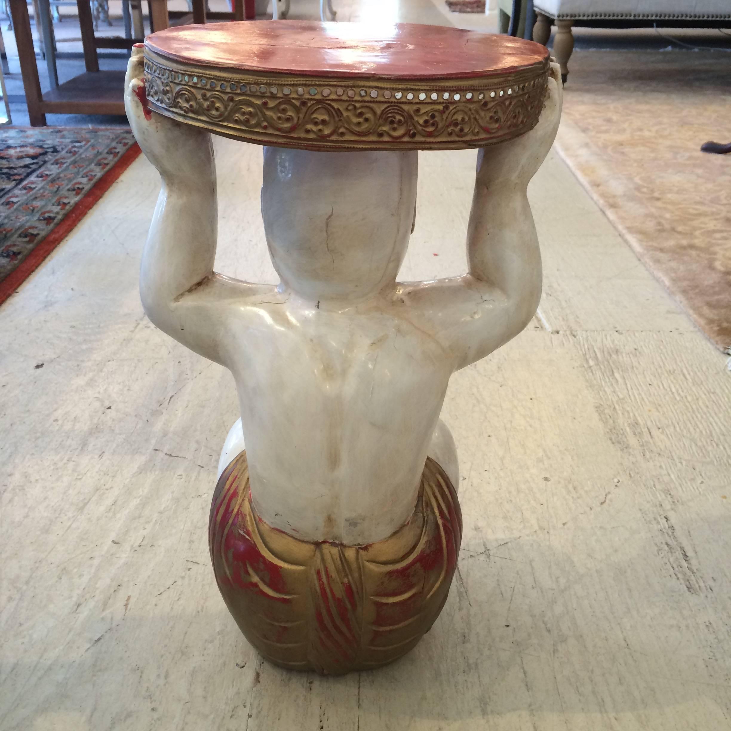 Fabulous little carved wood, painted and gilded drinks or side table in the shape of a Buddha holding the table surface on his head. Inset bits of mirror adorn the periphery of the table top which is a stylish Asian red.