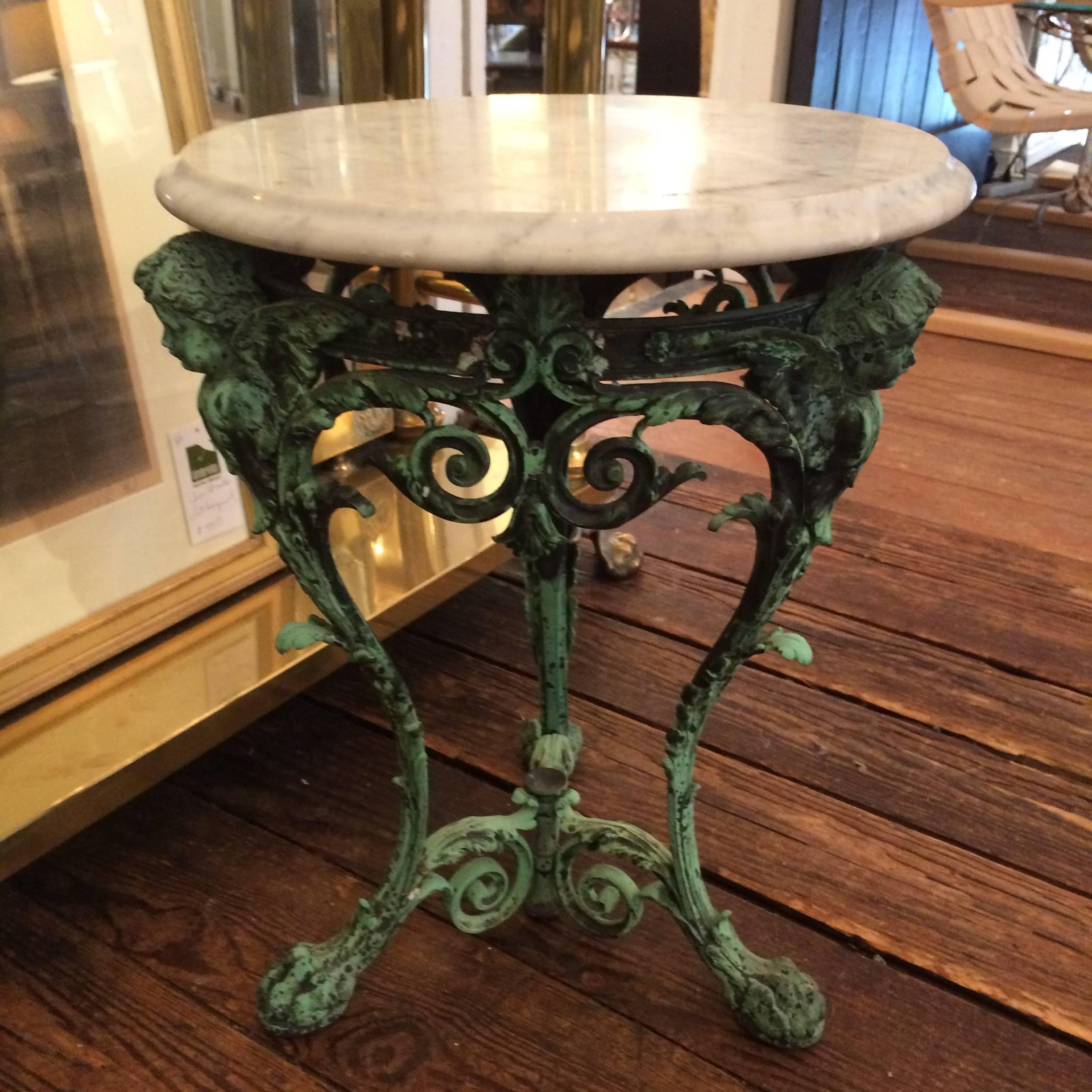 Gorgeous little side or drinks table with an ornate iron base with curlicues and putti, with an verdigris patina. A round white marble top rests on top.
