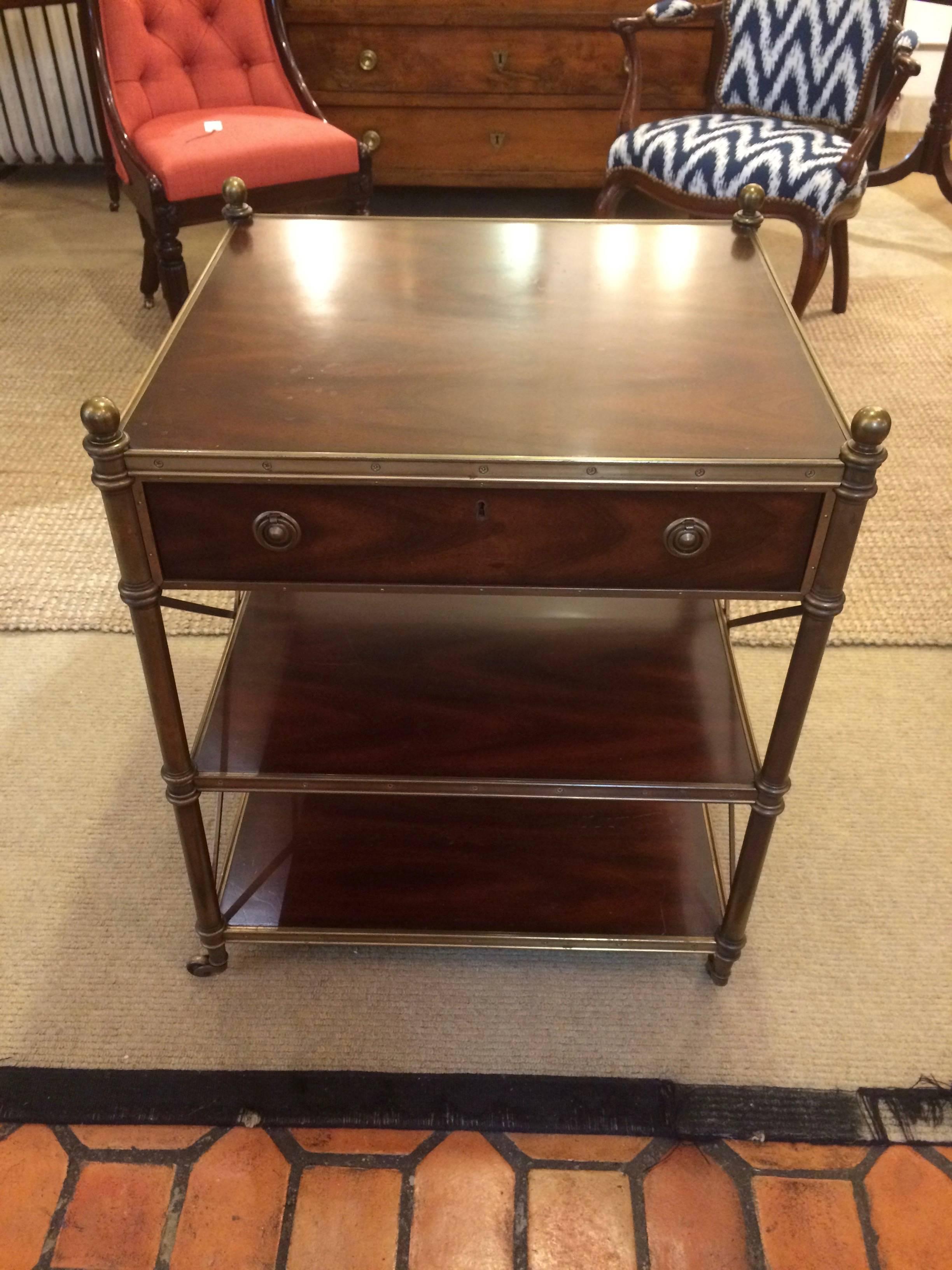 Campaign style side table or nightstand having three solid tiers of gorgeous mahogany and handsome brass with stunning visible screws, casters, key, finials and "X" stretchers on the sides as well as a single drawer. Measures: 28" H