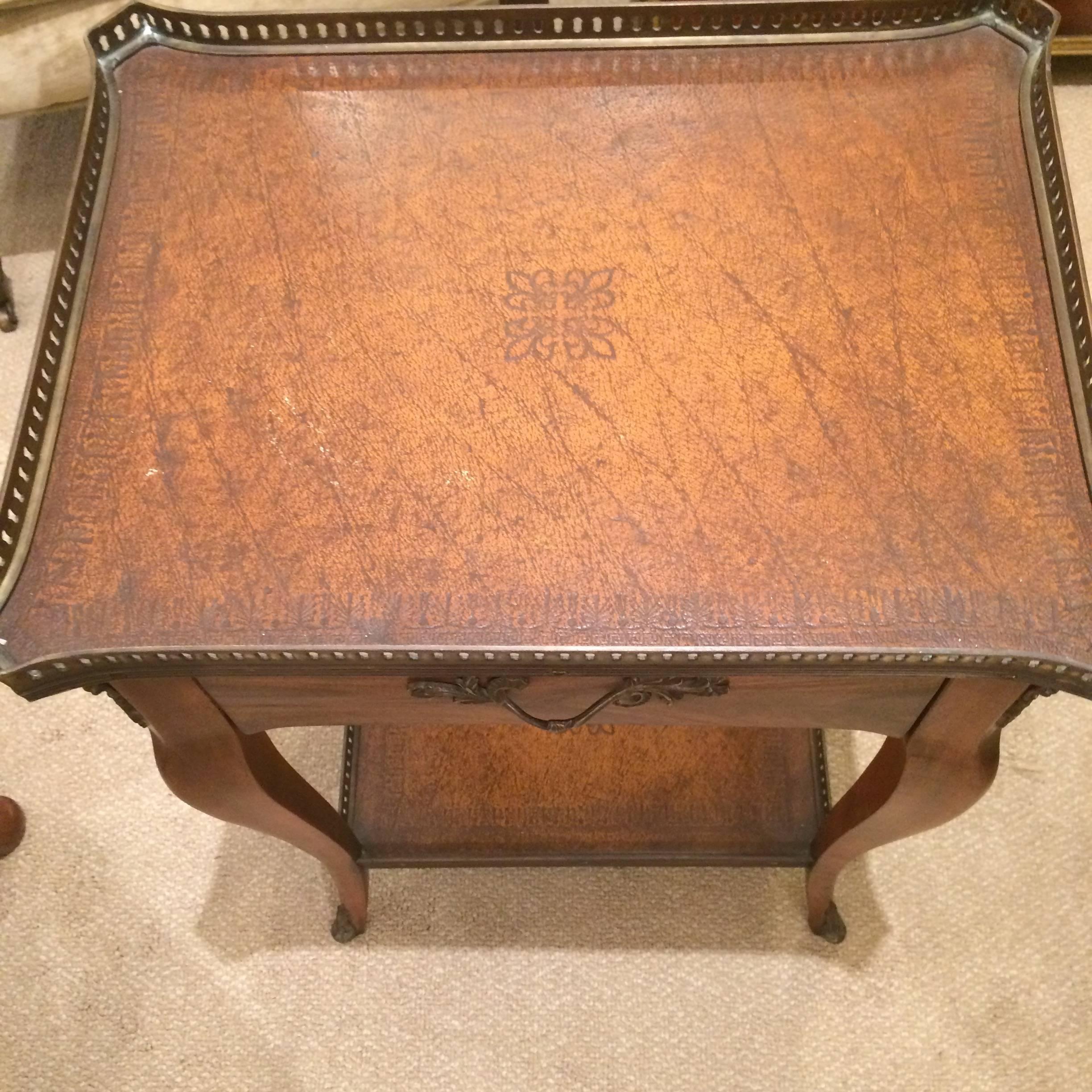 Handsome little traditional mahogany side table with two tooled leather tiers, bordered by brass galleries, with elegant curved legs with capped feet and a fancy curlicue handle on the single drawer.
