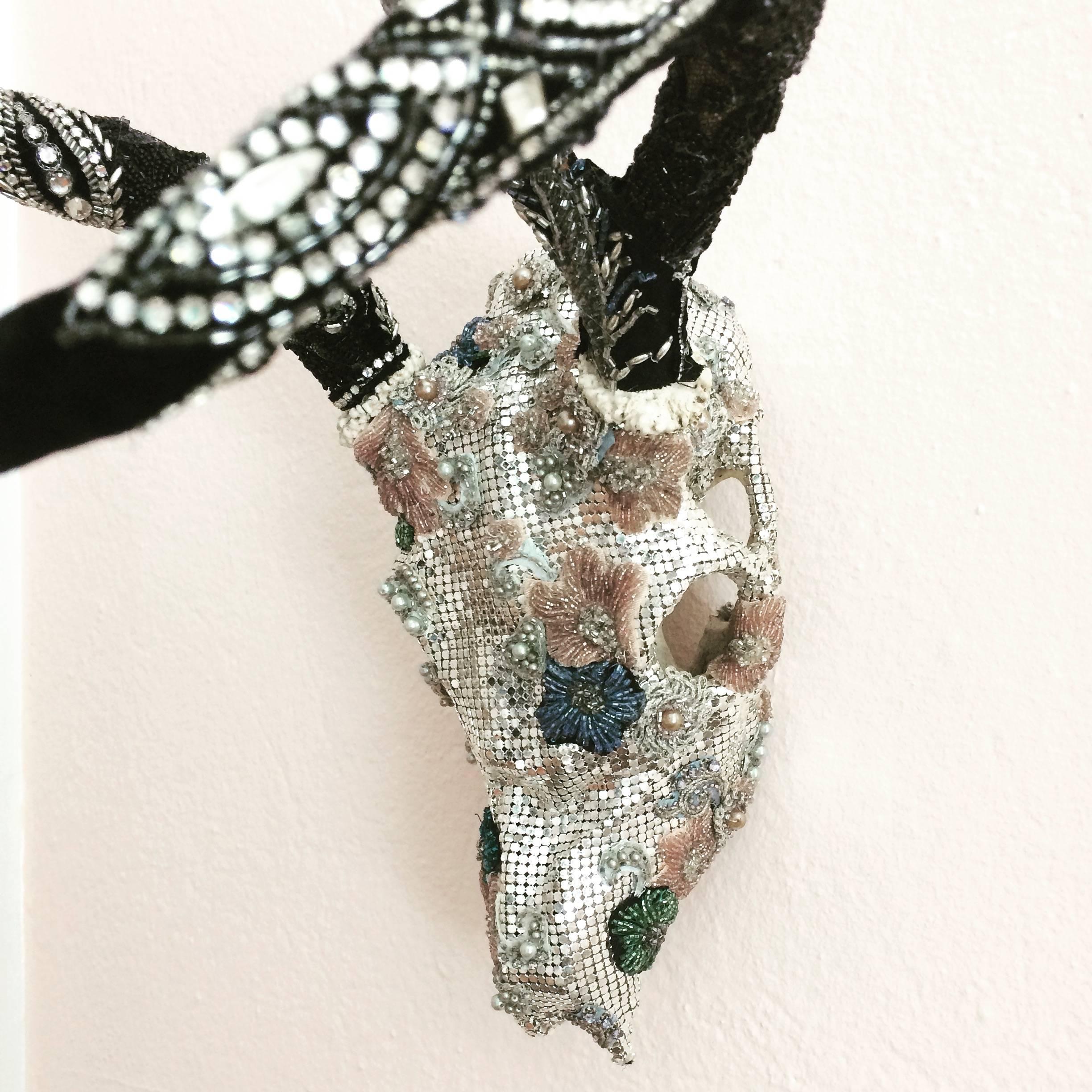 Una joya única de escultura mural de técnica mixta hecha con una pequeña calavera, meticulosamente adornada con piezas vintage de telas de cuentas, encaje y un bolso de noche de malla plateada.

Por Fay Sciarra.