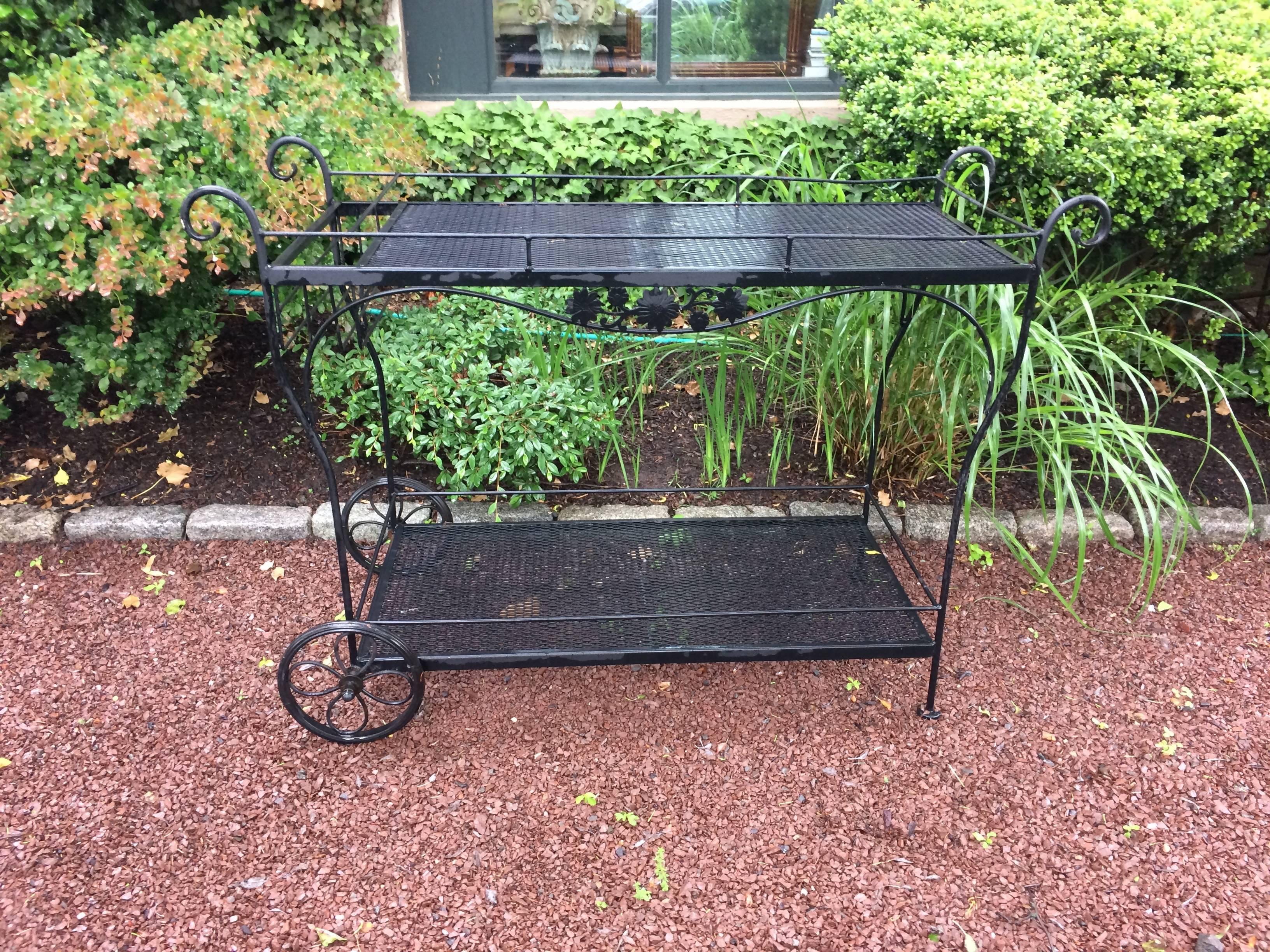 Classic black iron vintage Woodard bar cart having two tiers, a basket at the handle for liquor bottles and such, pretty wheels with daisy motife decoration within, and especially large size for entertaining at garden and POOL parties.