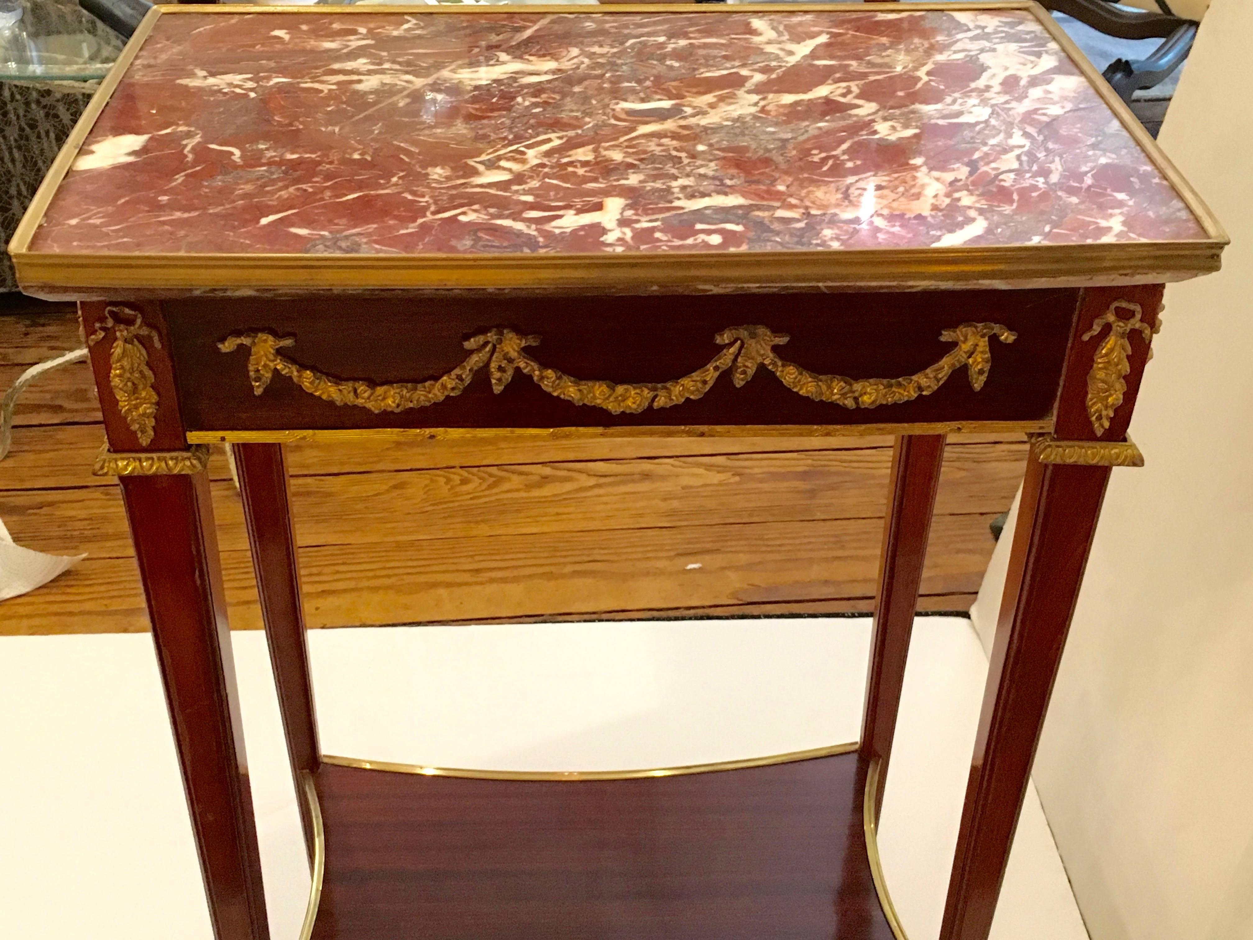 Lovely elongated French style end or side table having thin lovely legs, a second shelf below, cranberry and cream marble top with brass gallery surround and capped feet, topped off with pretty ormalu embellishments.