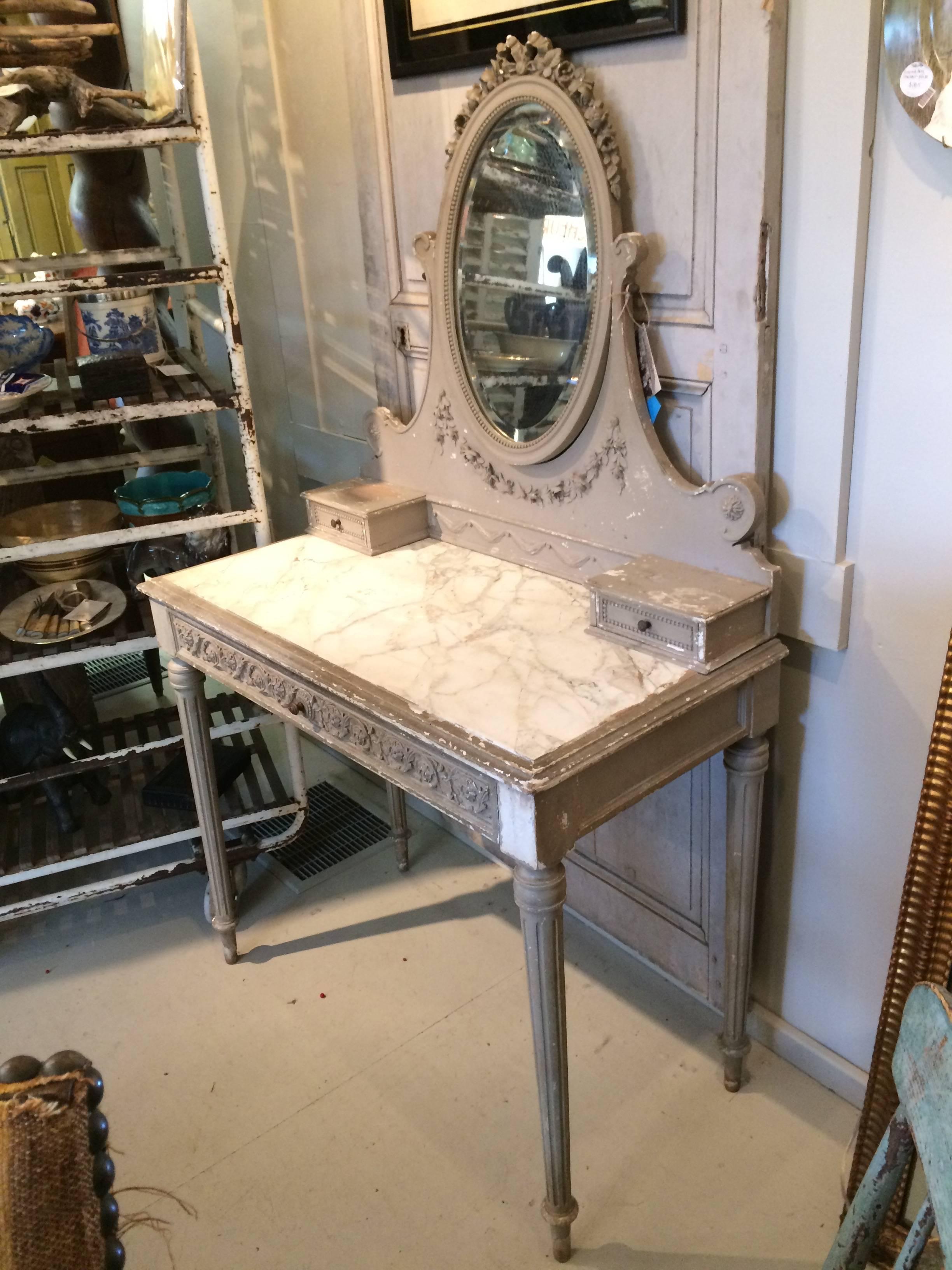 Gorgeous soft grey painted and carved wood French vanity having a lovely oval mirror with two small drawers at the base, a white and grey veined marble surface on top of a long single drawer, all sitting on top of tapered reeded legs.

In two pieces