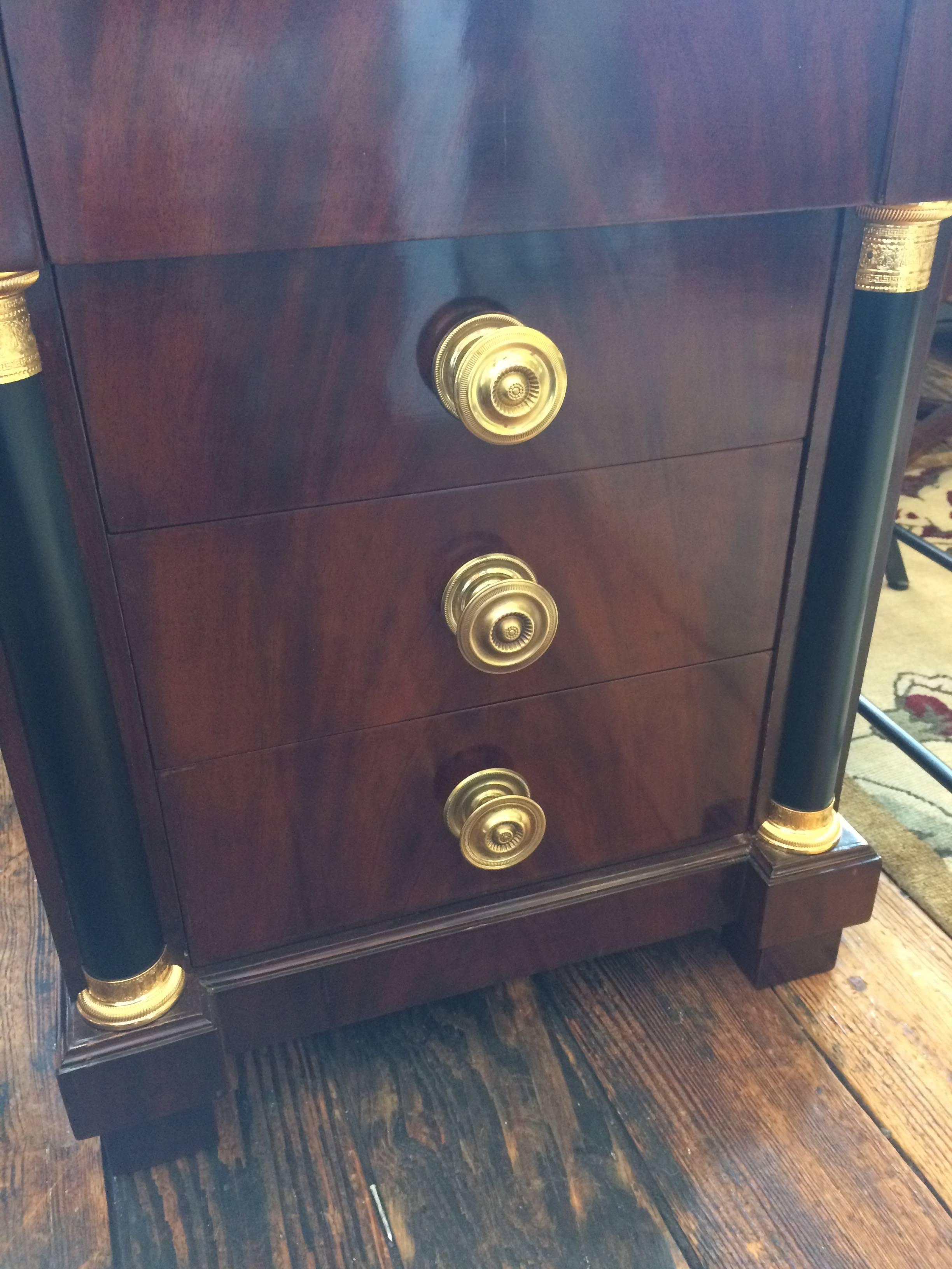 Handsome pair of mahogany nightstands or small bachelor chests having four drawers, the top drawer recessed so without a knob, brass hardware and details, ebonized columns on the fascade, and white marble tops.