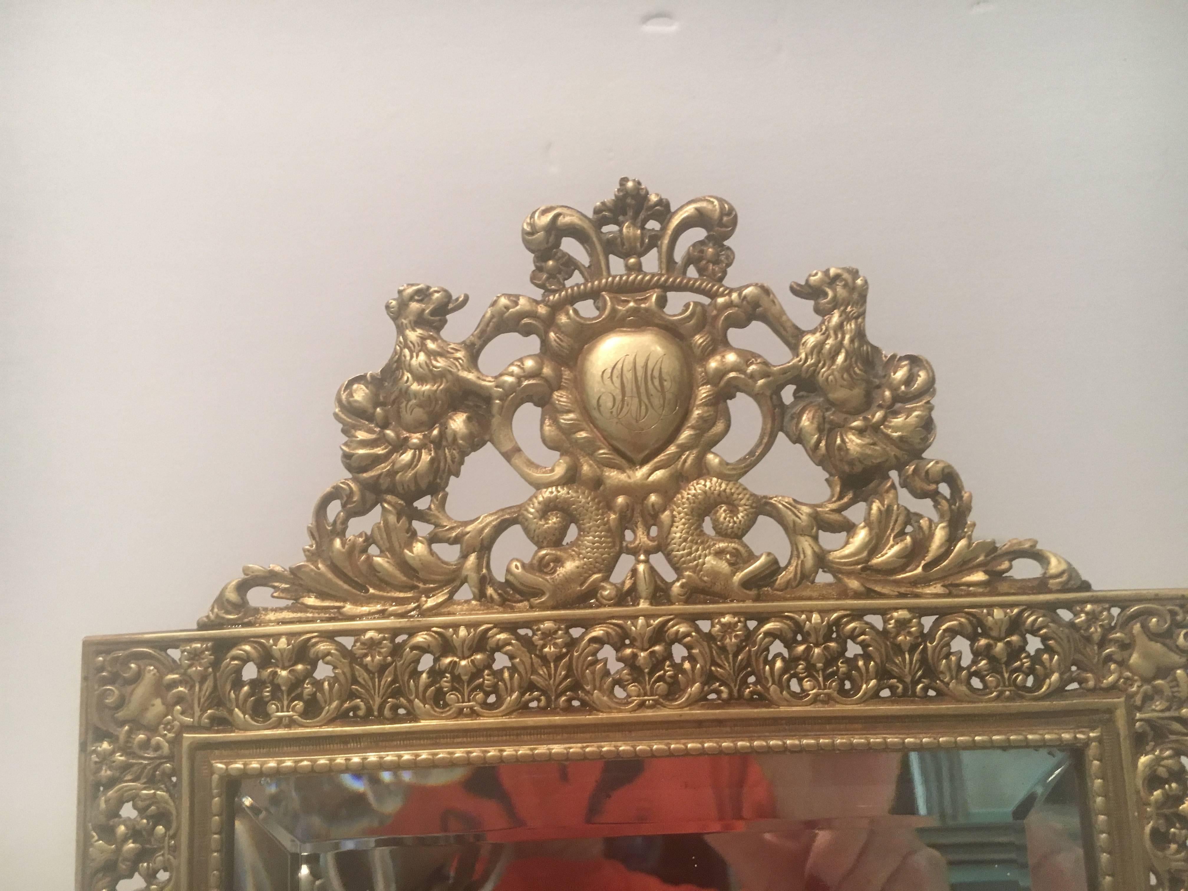 Stunning antique English dressing/vanity mirror in beautifully detailed gold toned brass. The monogrammed crest is encased in a heart surrounded by scrolled lions and dolphins. There is a loop on reverse for hanging on a wall.
     