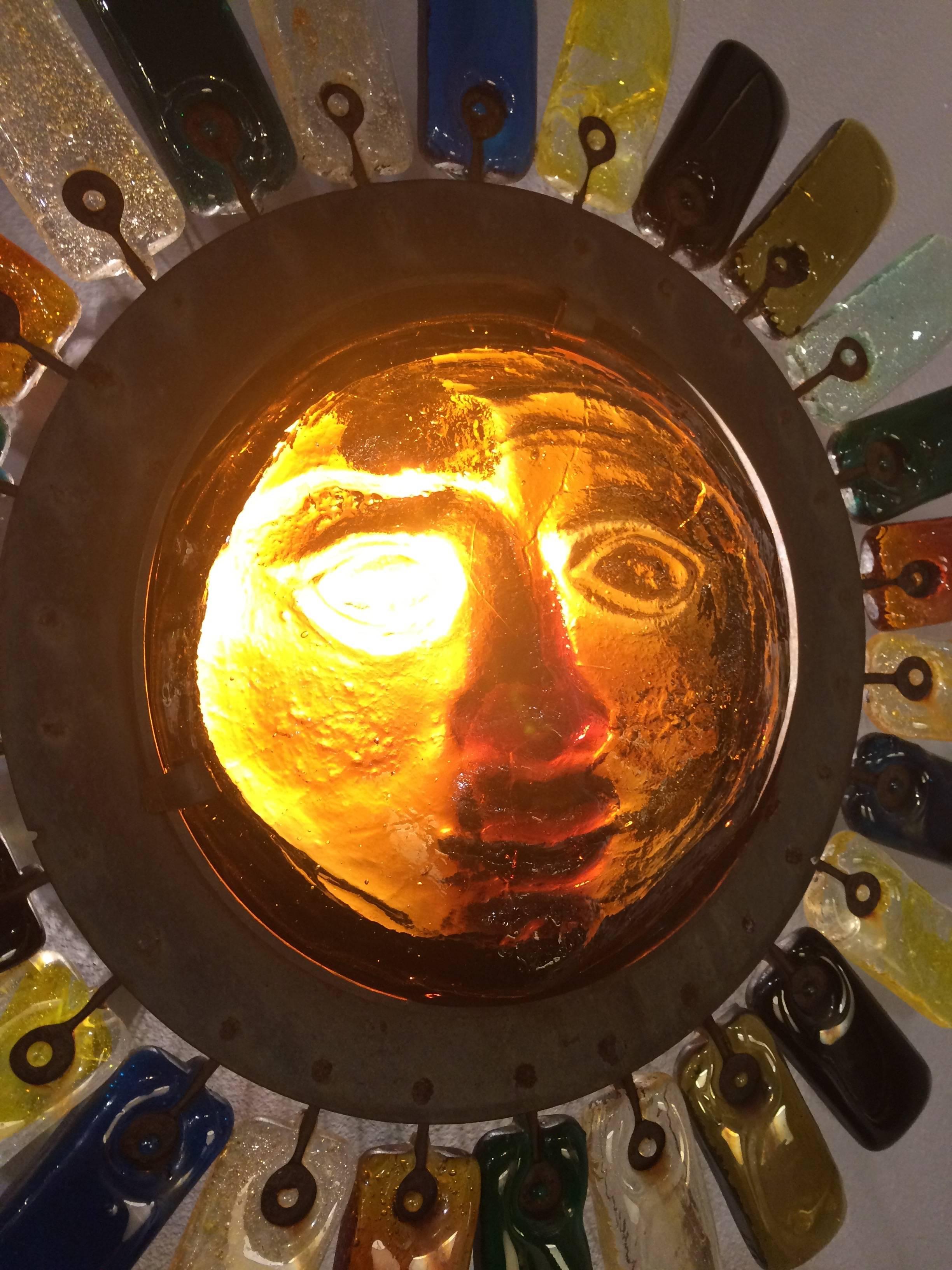 One of a kind handmade wall sculpture or sconce in the shape of a sun with a face that illuminates and multi colored glass 
