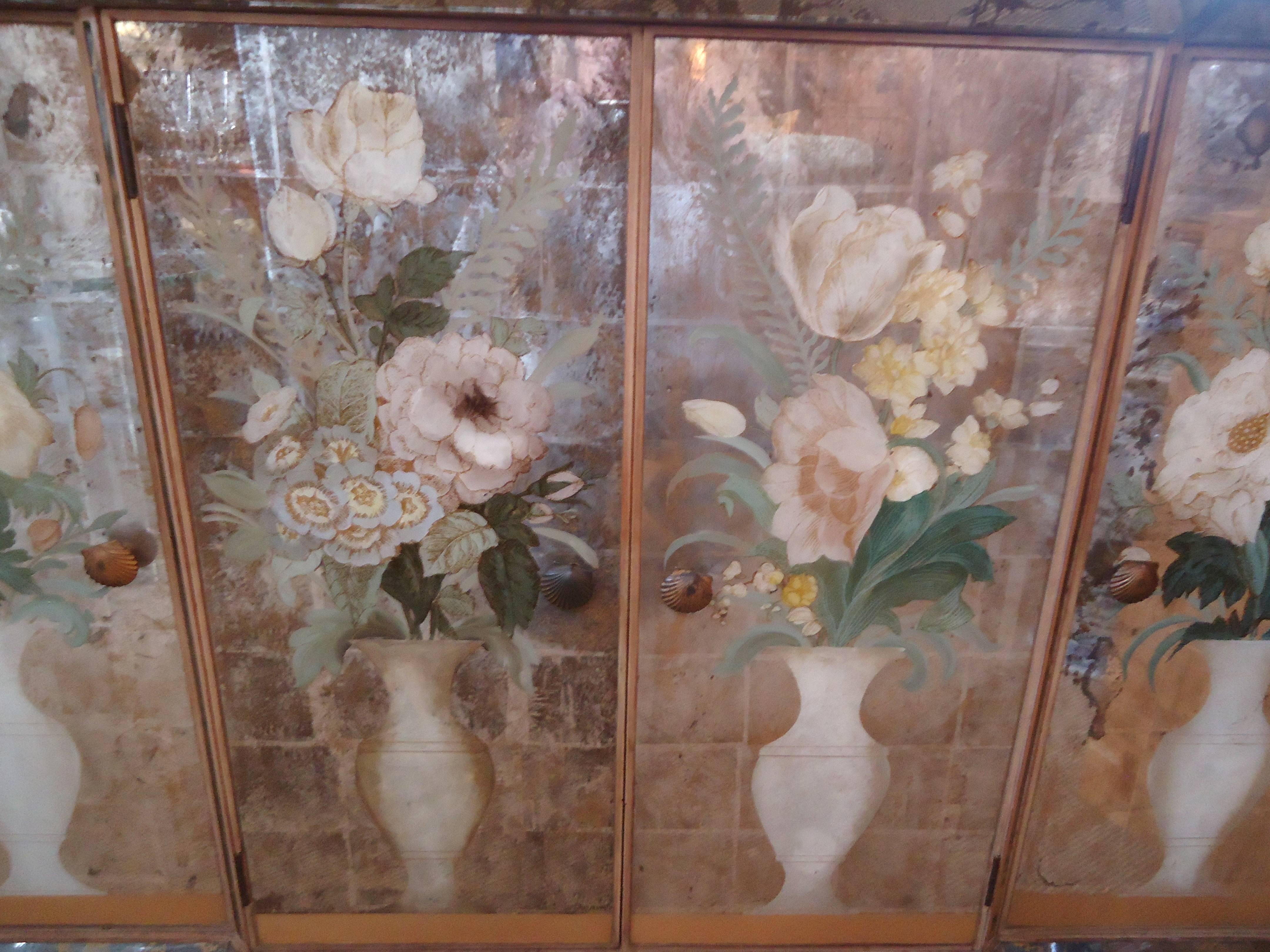 Magnificent reverse painting on glass (églomisé) of four white vases with lovely pale floral bouquets, silver leaf and mirror add glitz and glamour to this narrow credenza. Four doors with lots of storage within. Lovely bronze knobs in the shape of