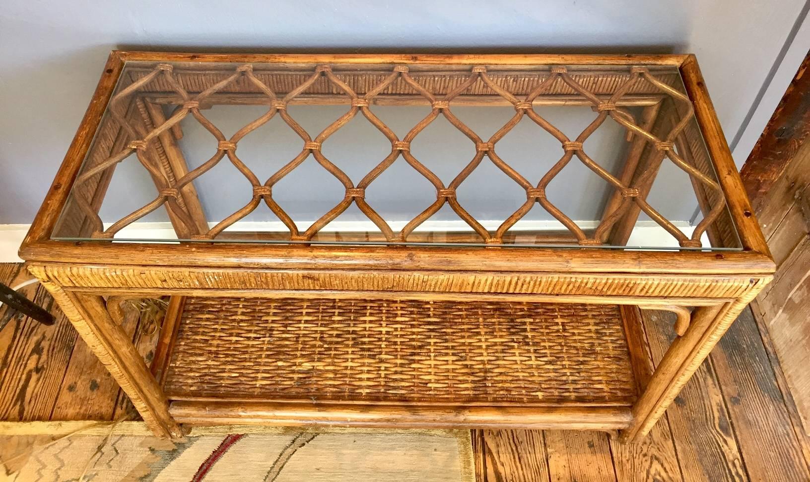 Stylish rattan console in a fairly short length, having lower woven shelf and top criss cross design seen through a piece of glass.