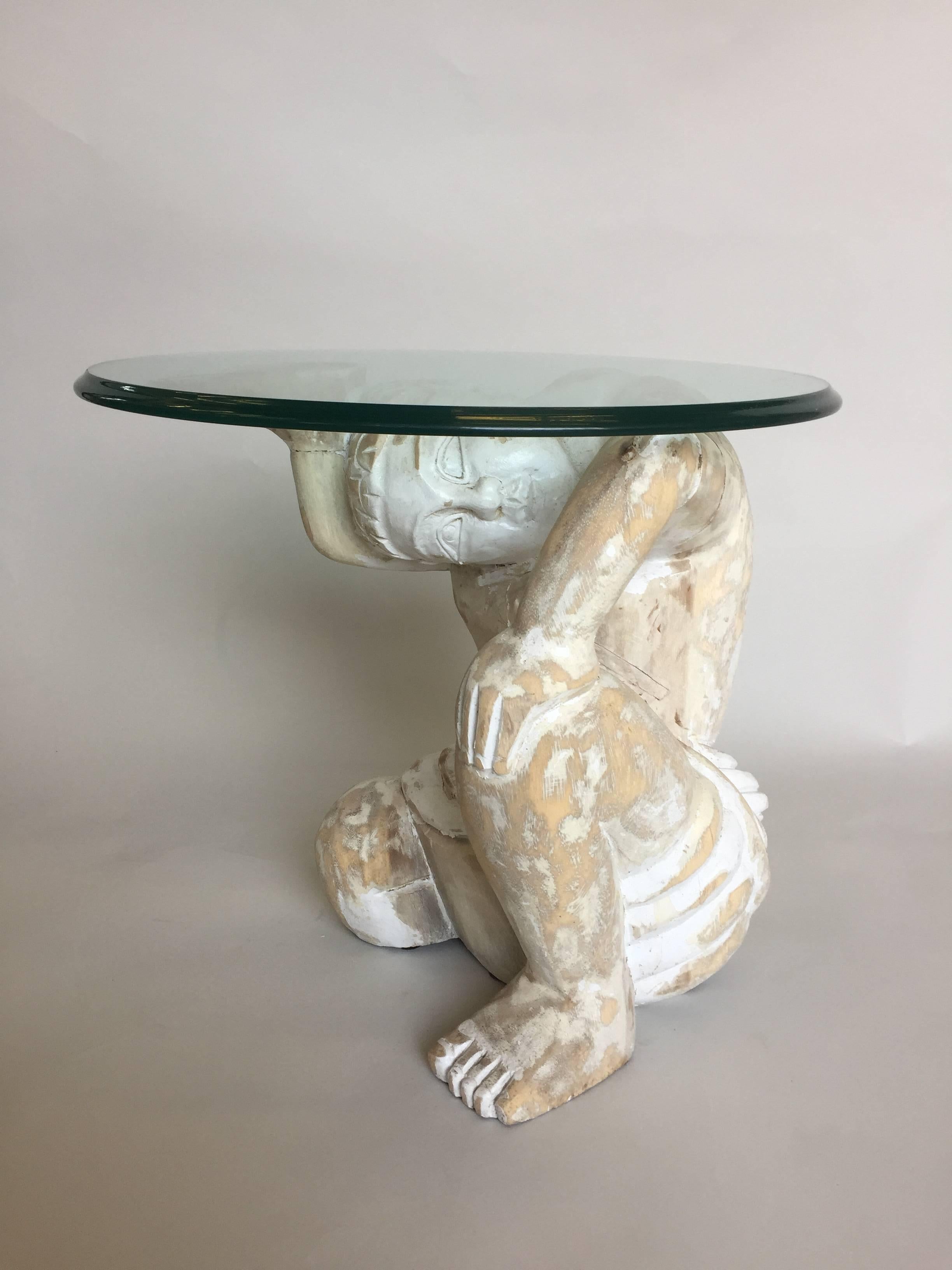 One of a kind little drinks table with big focal point value, having a chippy painted wood natural white and cream sculptural base of a muscular Asian warrior who holds up a 1/2