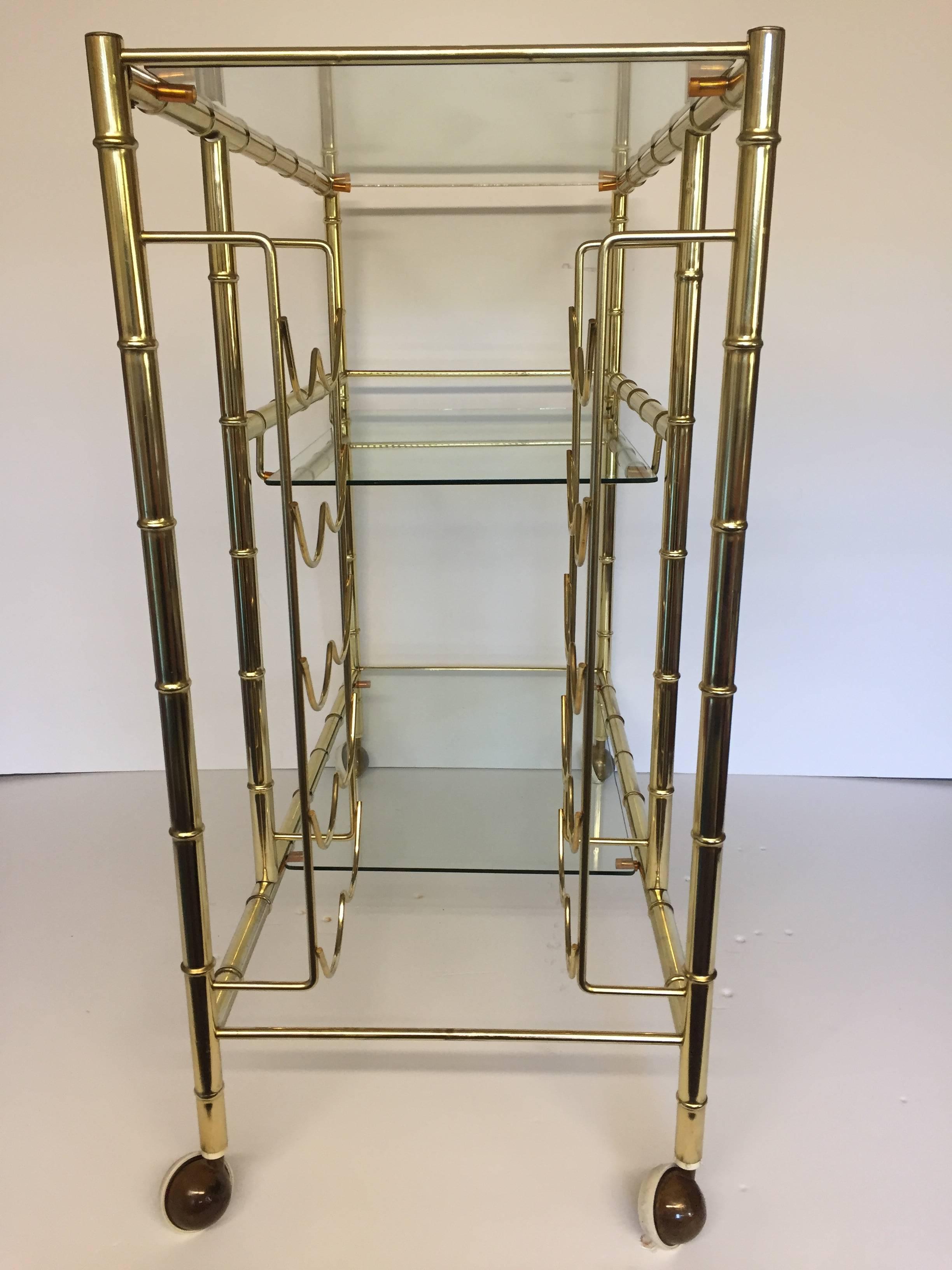 Stylish bar cart with a sophisticated unusual design having a square shape with three glass shelves and a bottle or wine rack to one side. The brass-plated body has a faux bamboo look.
 
