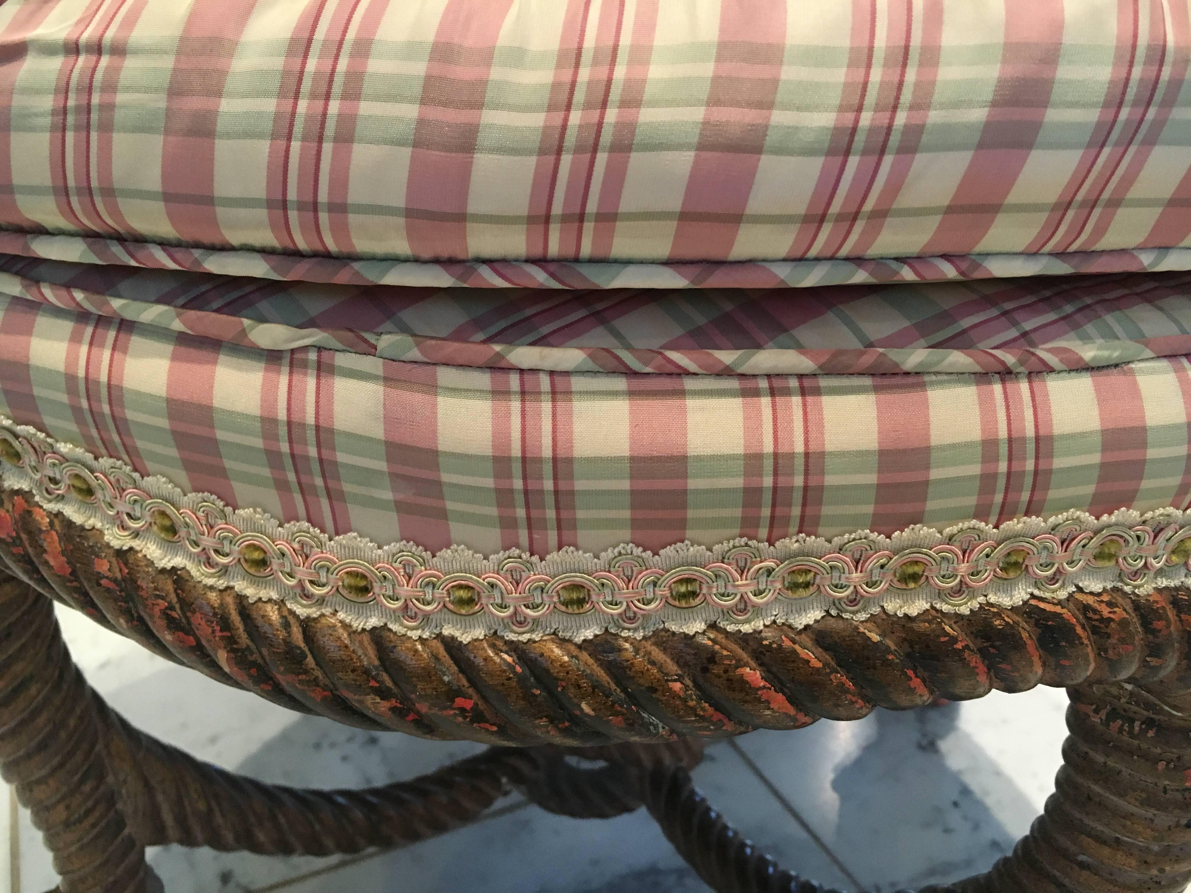 A carved partially giltwood wood ottoman or stool in the form of a knotted rope. A salmon color is visible where the gilt has been worn, only adding to its character. Shown in a small silk plaid of rose and celery with a top false tufted cushion.