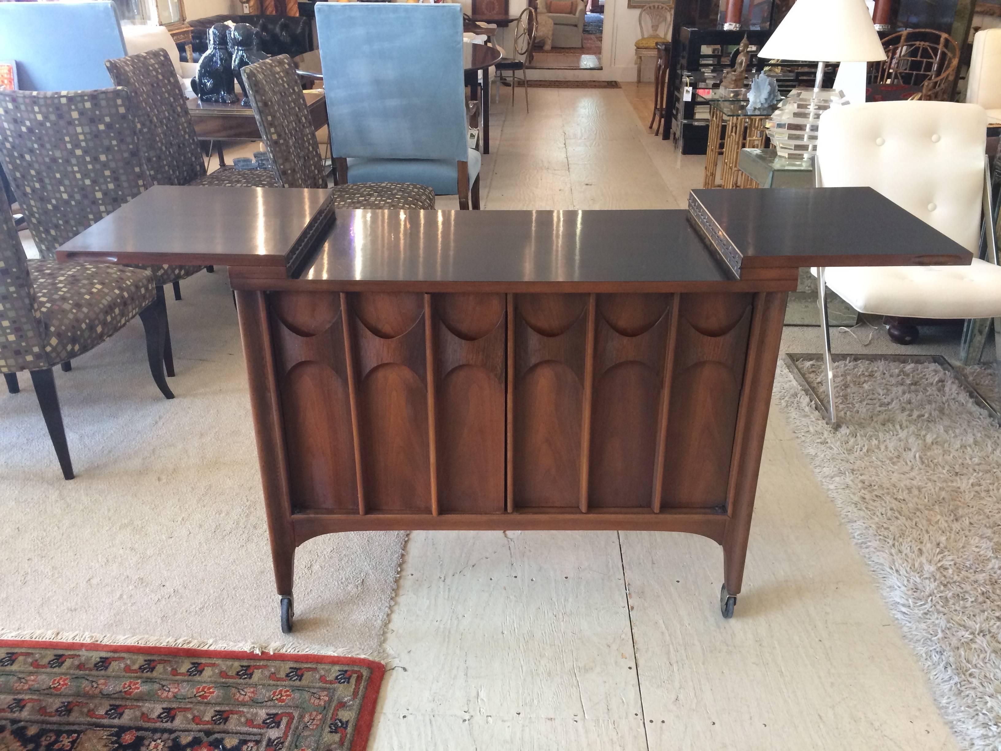 Beautiful mahogany server or bar cart with Danish modern vibe, sculpted 3 dimensional front doors that open to storage within. Top opens out to reveal a black formica surface. Tapered feet on casters. Fully extended 65.5 W.