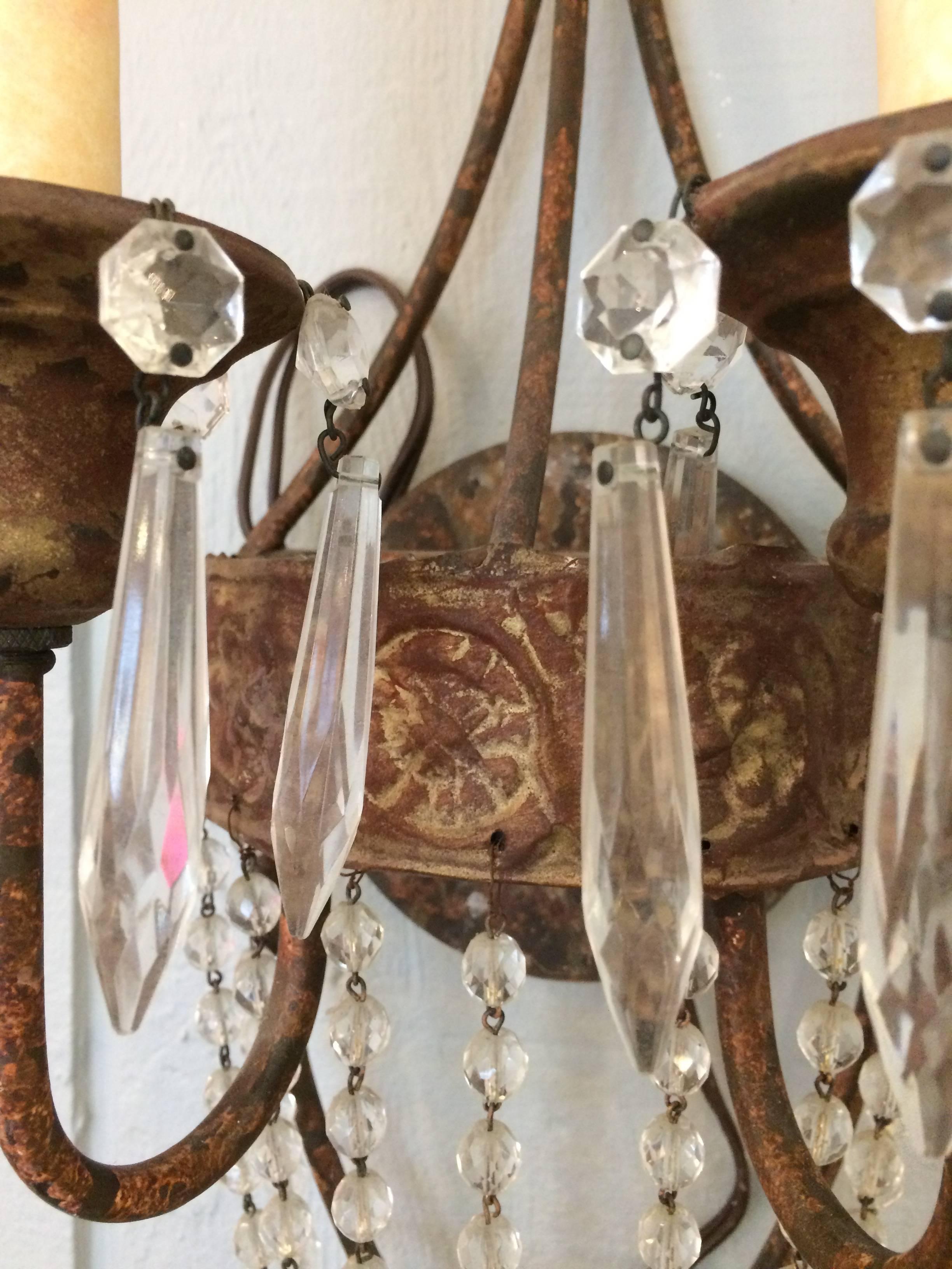 Two iron and tole wall sconces having two arms and a bronze gilt patina as well as lovely cascades of crystals with a jewel of a round prism at the bottom.

Note: There are seven sconces available.