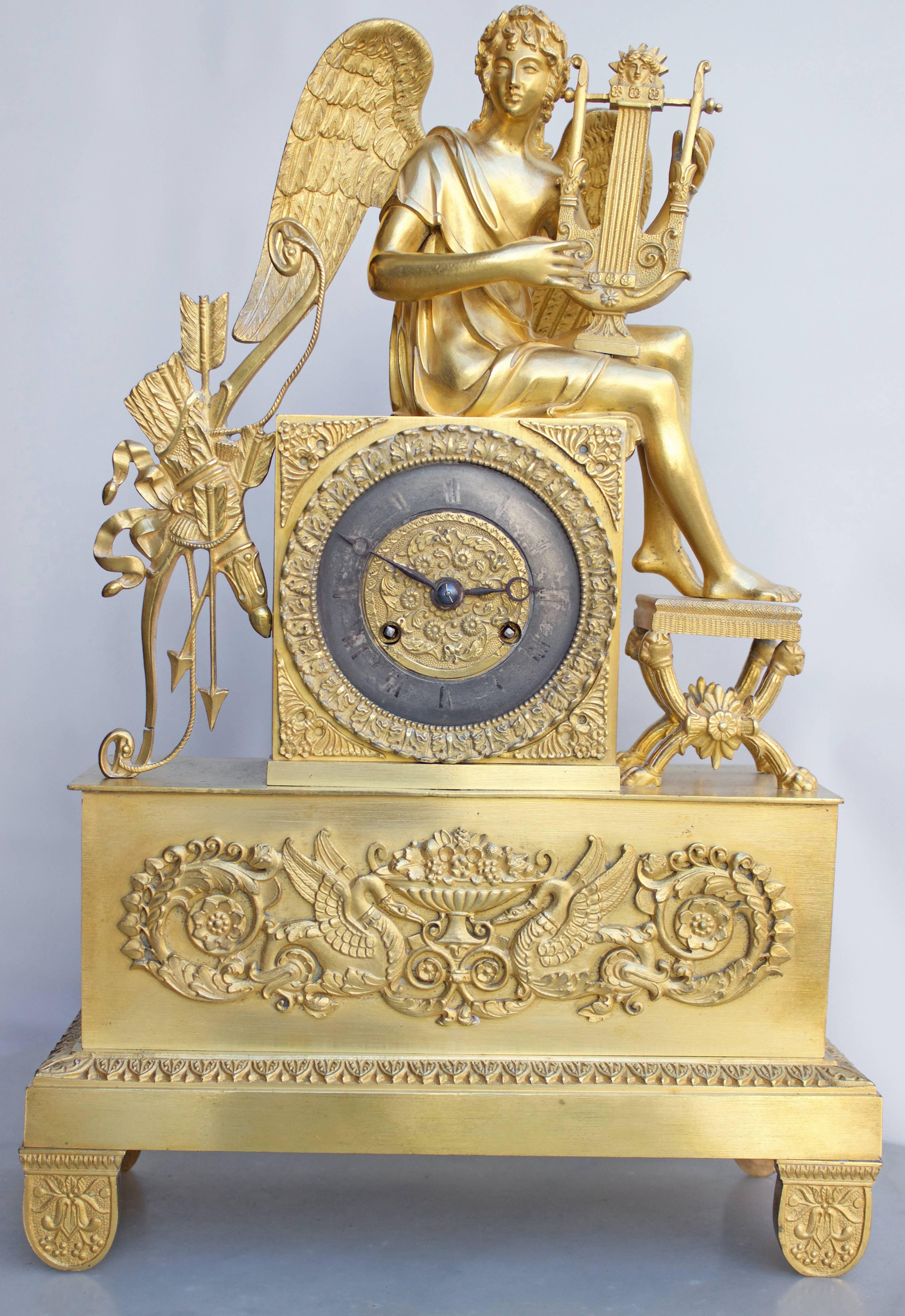 At top, a winged teenaged Angel dressed a L'Antique holding a lyre and resting his feet on a curule shaped seat. On left a quiver with its bow and three arrows. The clock has a blackened steel dial with engraved Roman numerals and hand cutted iron