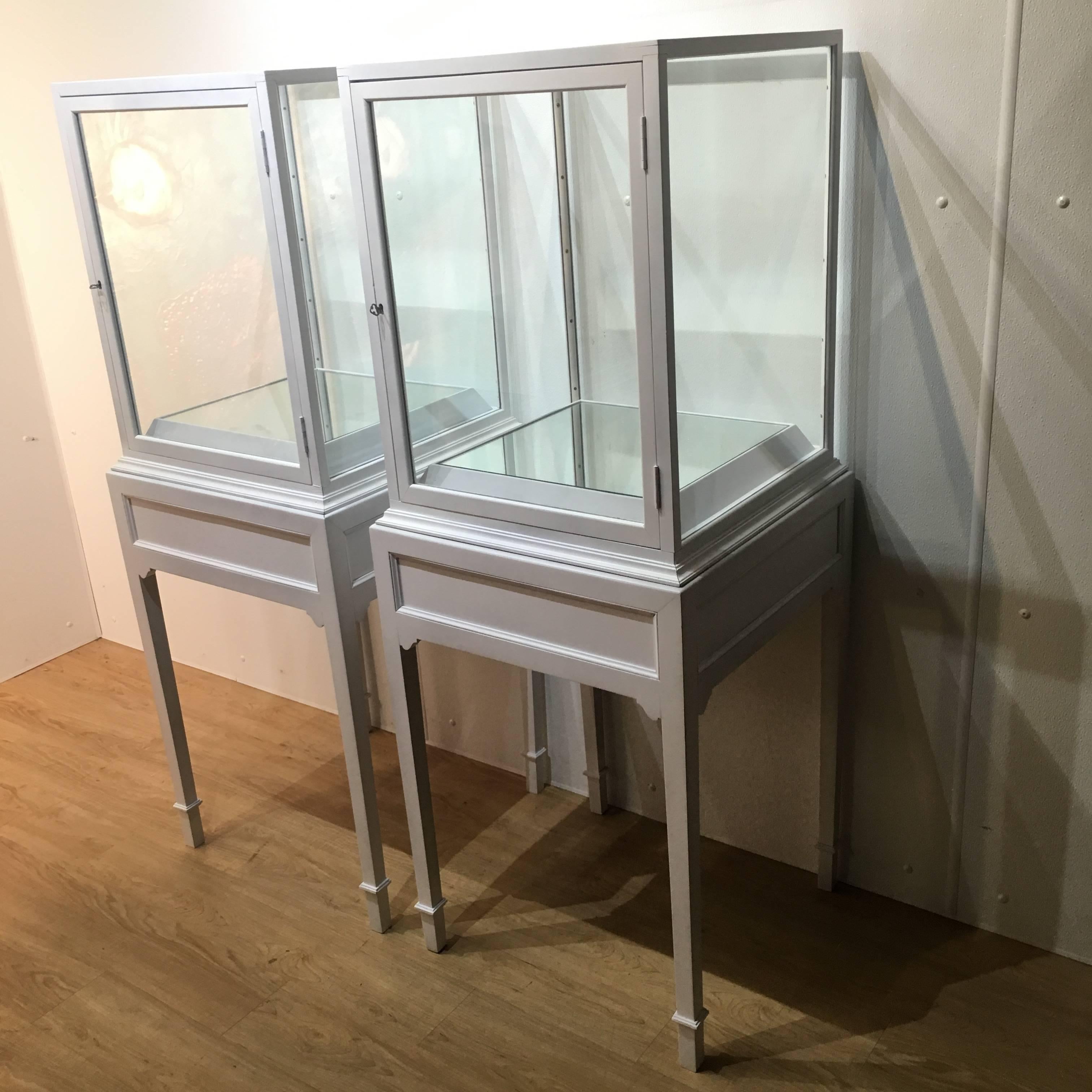 A pair of white lacquered display cabinets. Each one fitted with all around glazed panels, one as a door. The interior has 3" high mirrored plinth/pedestal. Raised on four tapering legs. Complete with working locks and keys. Two piece