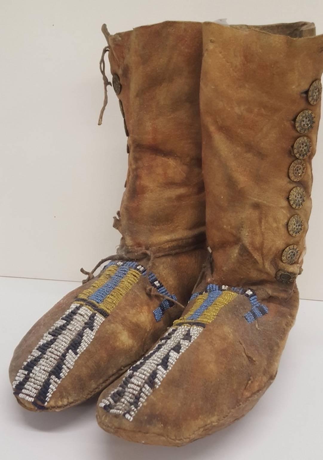 A pair of high button, deerskin moccasins of a Plains Native American tribe.
Beaded by hand on each moccasin top, in yellow and blue above a black and white saw tooth band or strip of period beads, down to the toe.
There is a row of machine made