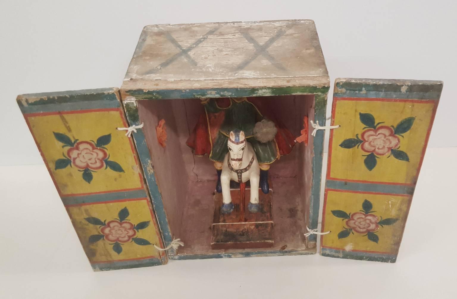 A small Bulto of Santiago Matamoros mounted on his customary white horse, set inside a soft-pink painted wood box (portable shrine) with charming, pink paper flowers and Primitive, floral painted doors on string hinges. When closed the wooden box is