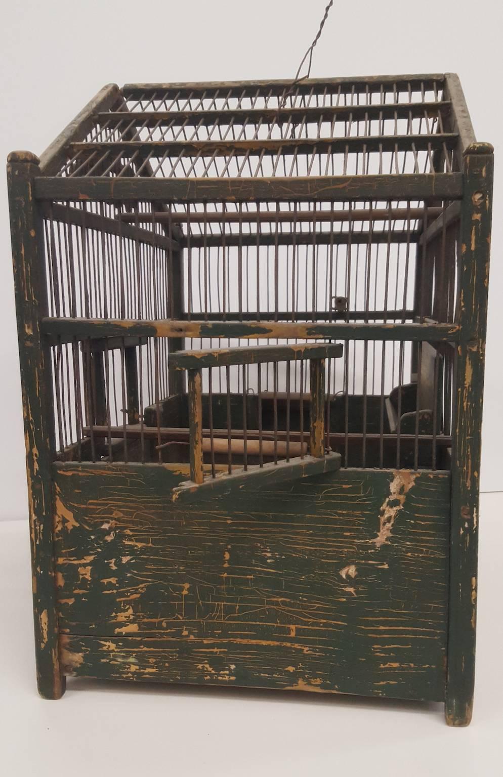This good sized French bird cage is painted a dark green. Over time the paint has become heavily distressed, showing a mustard paint beneath the green, with occasional chips, cracks, and losses showing the bare wood below. It has multiple perches, a