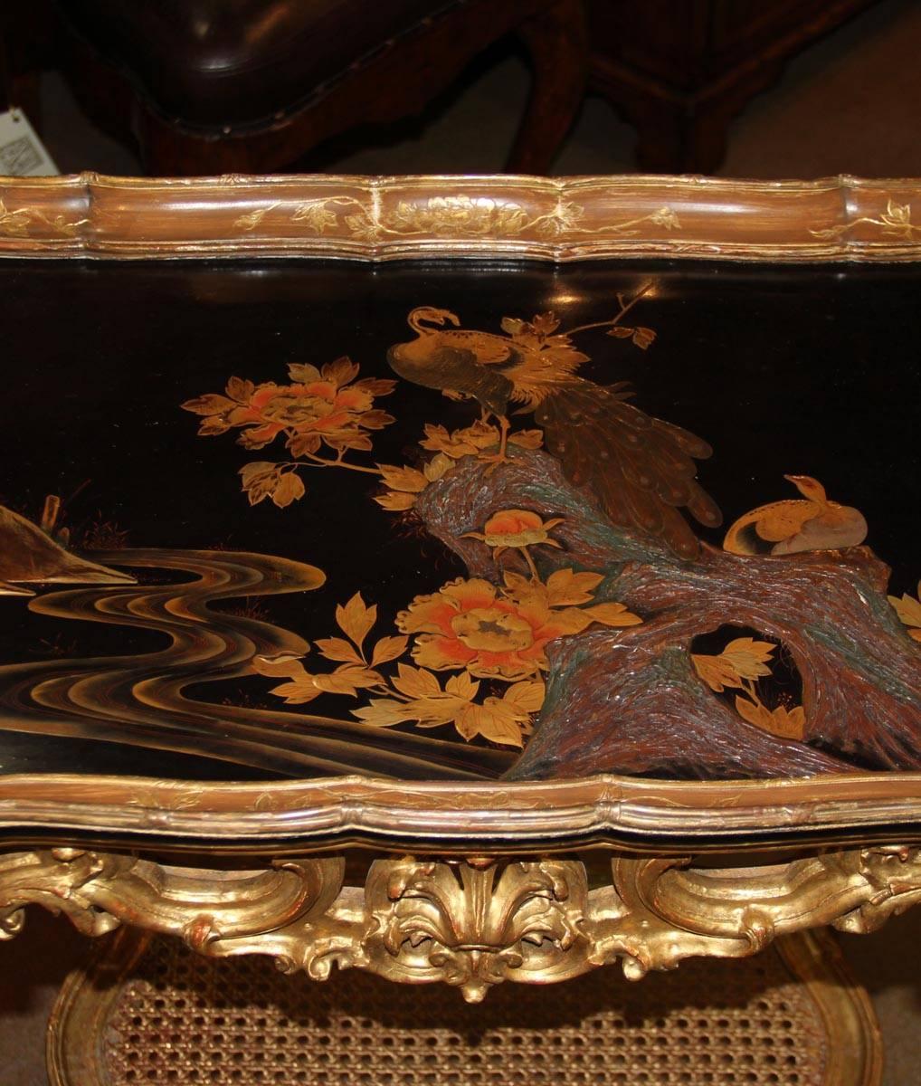A 19th Century French Giltwood and Black Lacquered Japanned Table, the dished top featuring two beautiful peacocks (with iridescent feathers) among rocks and flowers, and with a fretted apron centered by a scallop shell above a caned and gilded