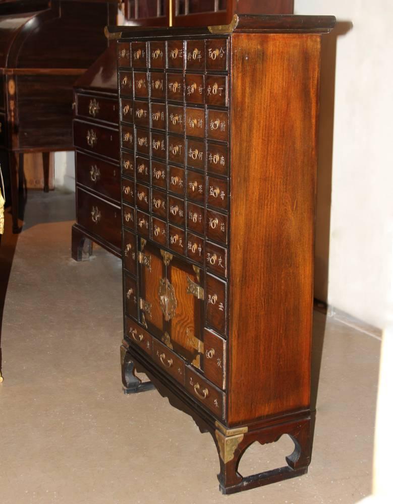A 19th century Korean rosewood apothecary chest with a torii-style top surmounting an array of 49 drawers, each carved with the name of a medicinal herb, as well as a pair of doors with a round latch and a fish-shaped lock and key (symbolizing