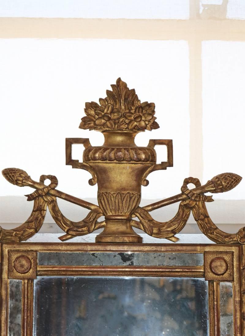 An 18th century French neoclassical Louis XVI giltwood mirror crested with an urn filled with flowers, flanked with lambrequins and thyrsus (in ancient Greece and Rome, a staff tipped with a pine cone and carried by Hermes and followers of