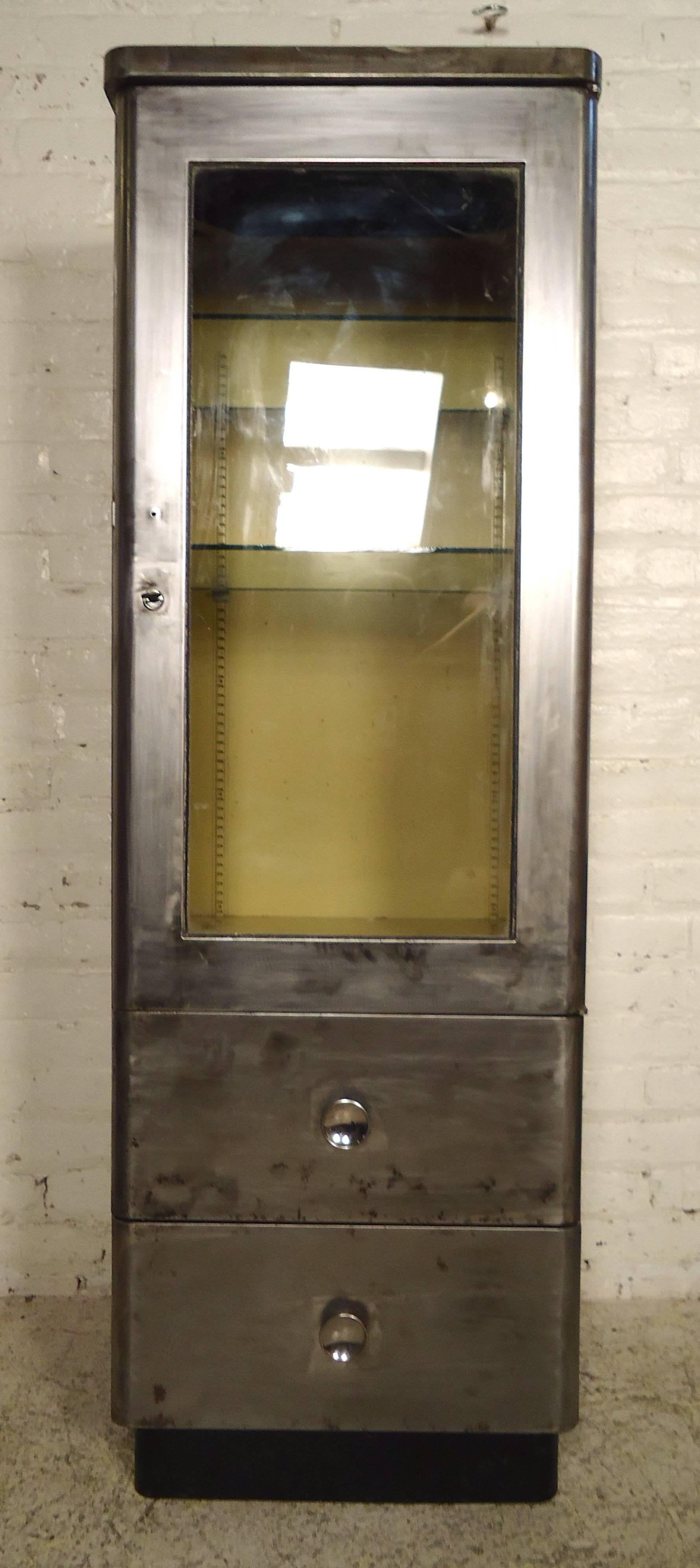 Vintage Industrial display cabinet featuring glass door cabinet with two adjustable glass shelves, with two lower drawers. Refinished in a bare metal finish. Key acts as door latch.

Please confirm item location NY or NJ with dealer.