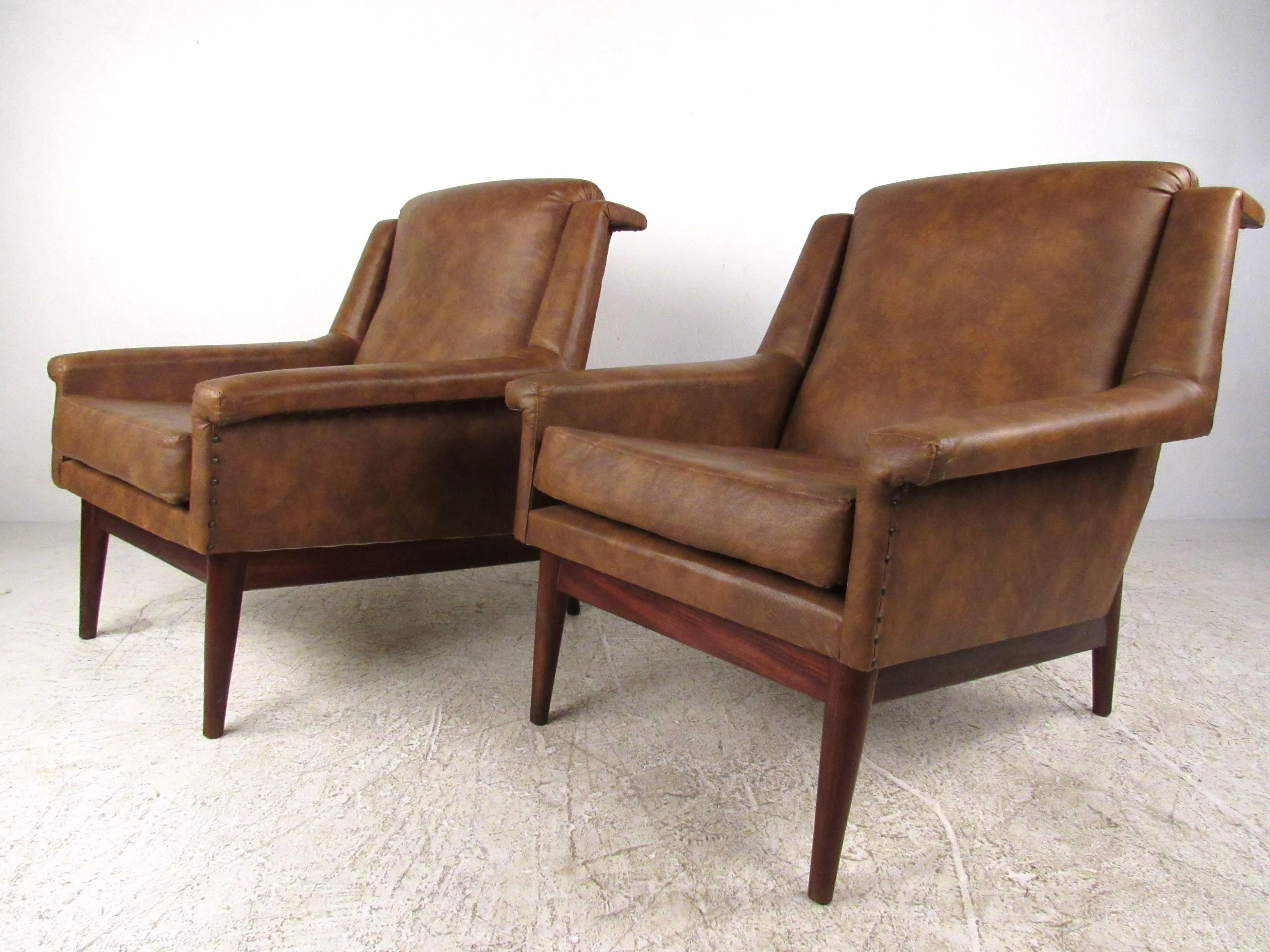 This unique pair of vintage vinyl lounge chairs feature wide upholstered seats with unique sculpted armrests. Vintage vinyl with brass studs adds to the classic appeal of the pair, while tapered hardwood legs showcase the Mid-Century influence on