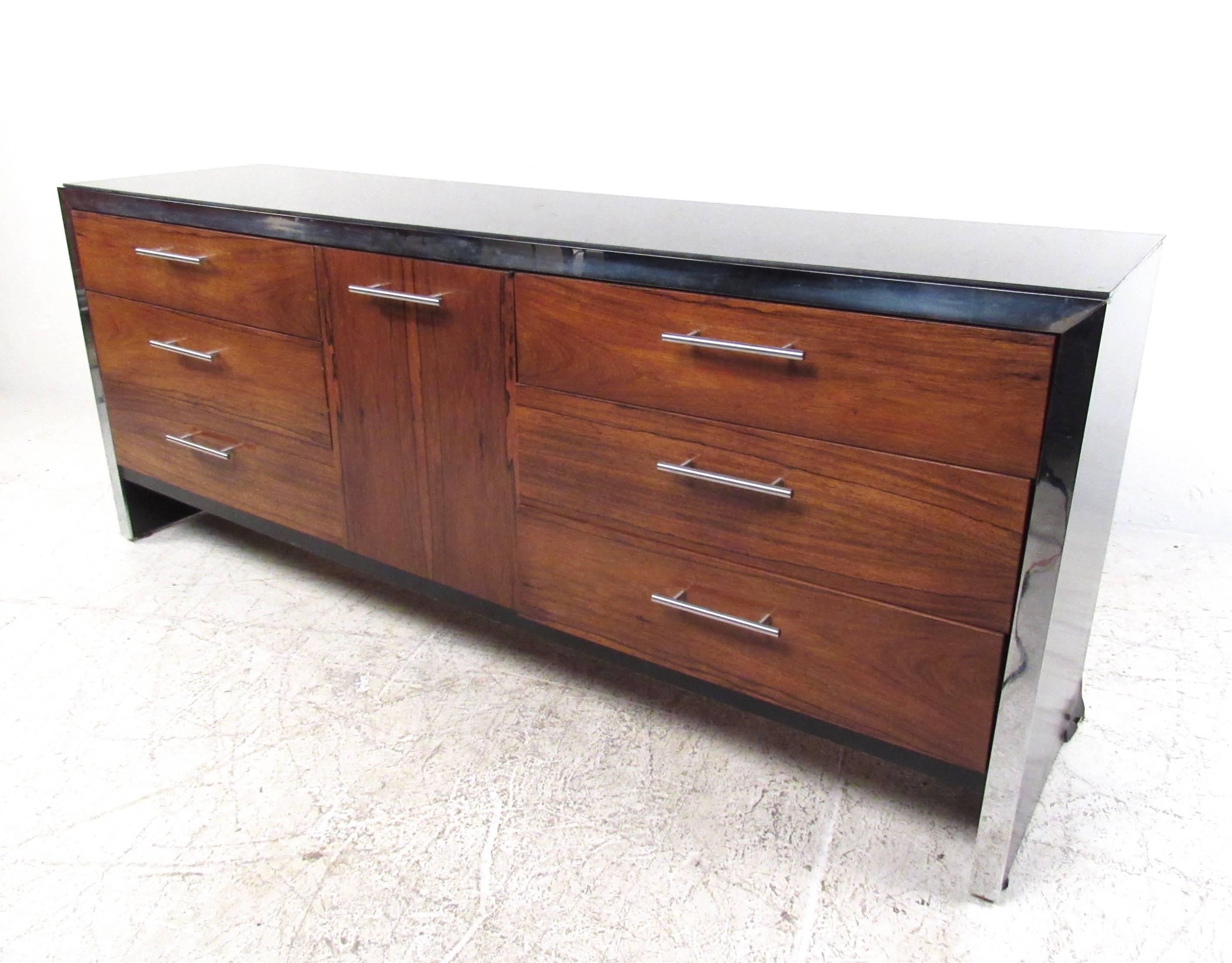 This stunning vintage dresser features stunning veneer front, chrome trim, and black glass top. Exquisite mid-century modern storage piece perfect for bedroom or other interior setting, this piece features the design of Milo Baughman as produced for