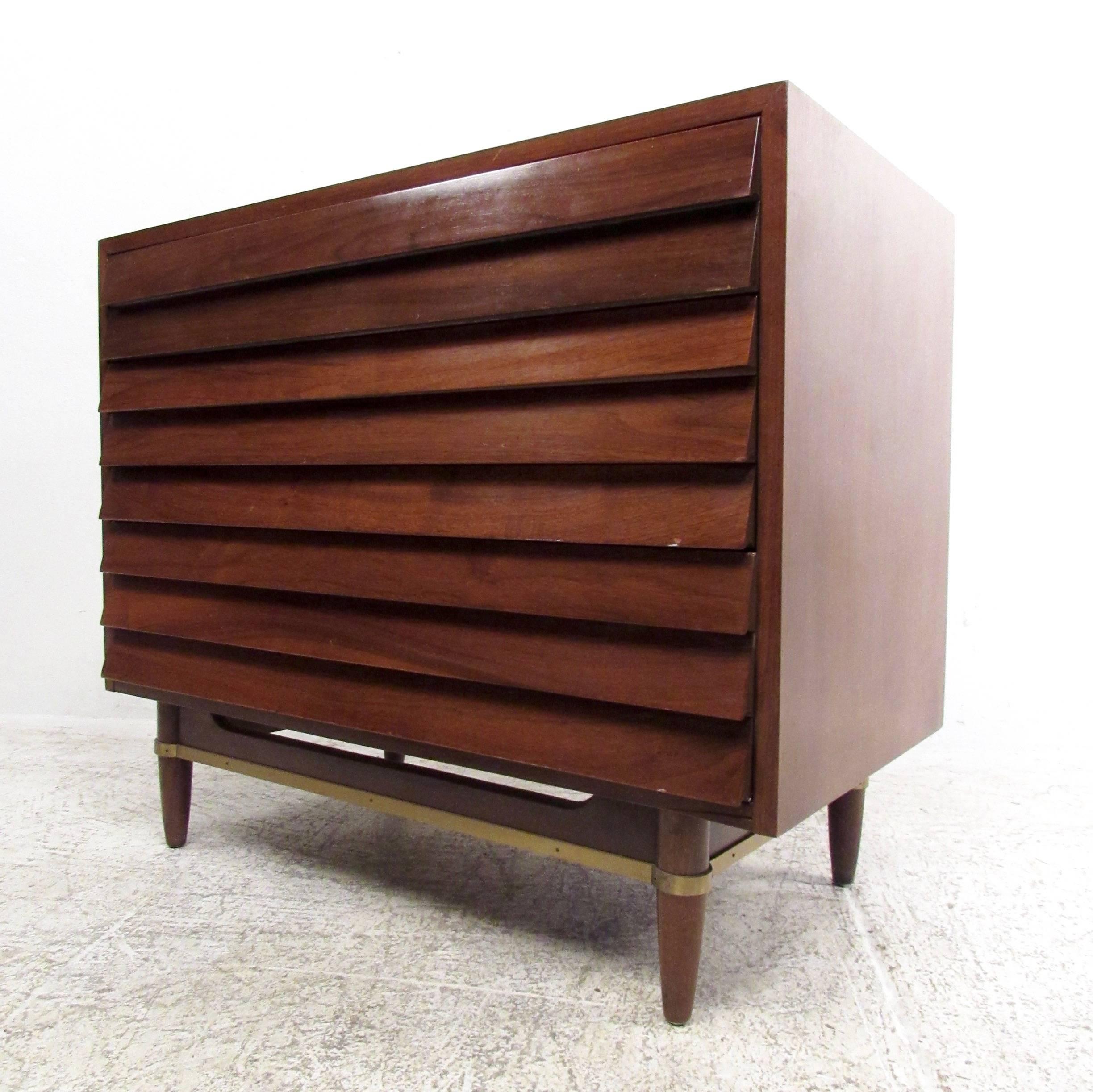 This matching pair of Mid-Century Modern sculpted front chests features three-drawer construction for plenty of storage with unique louvered front design. Vintage walnut finish and quality construction are accented by brass stretchers and drawer