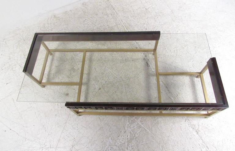 This unique designer coffee table features an offset glass tabletop mounted on a brass and carved wood base. Sturdy Mid-Century Modern construction mixes modern clean lines and ornately carved wooden trim. Please confirm item location (NY or NJ).