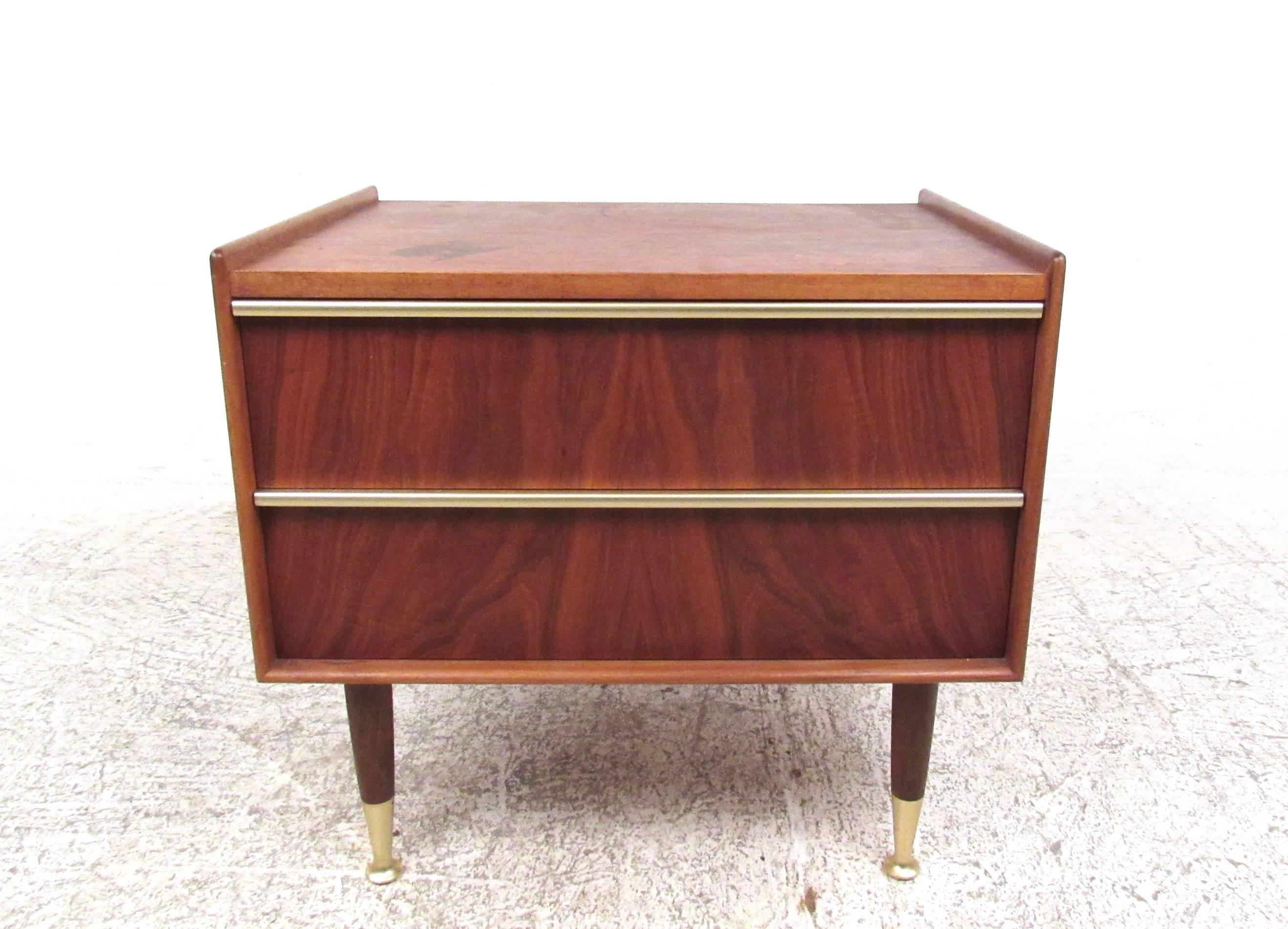 This single nightstand by Edmond Spence features unique raised edge design with brass finish trim on drawer pulls and feet. Tapered legs and rich natural finish add to the Mid-Century Modern style of this Swedish made piece. Two drawers for stylish