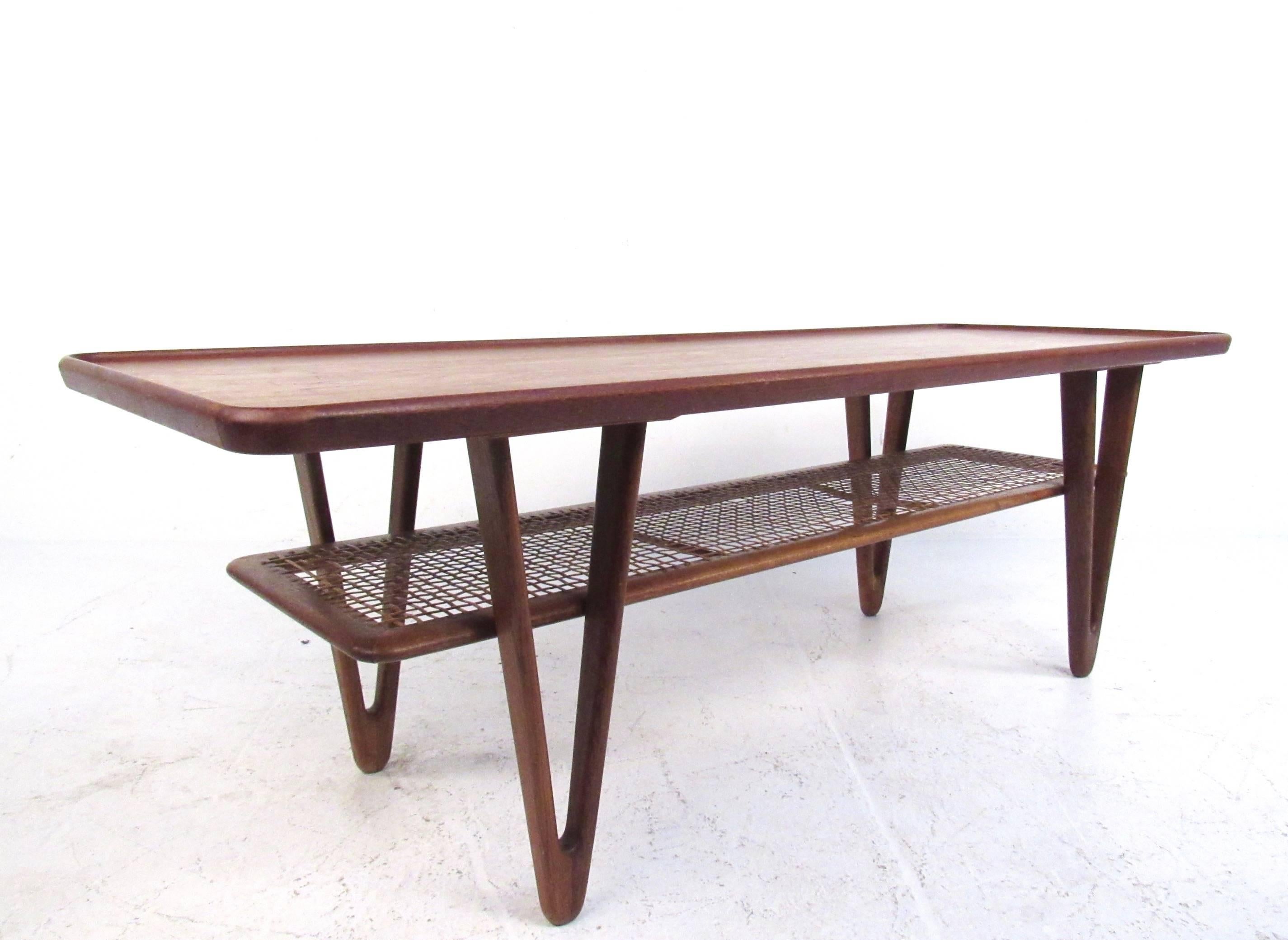 This stunning sculpted coffee table features unique triangular legs with second tier cane shelf. Raised edge teak top showcases the beautiful attention to detail paid by Kurt Ostervig in his iconic Mid-Century Modern designs. Stylish Mid-Century