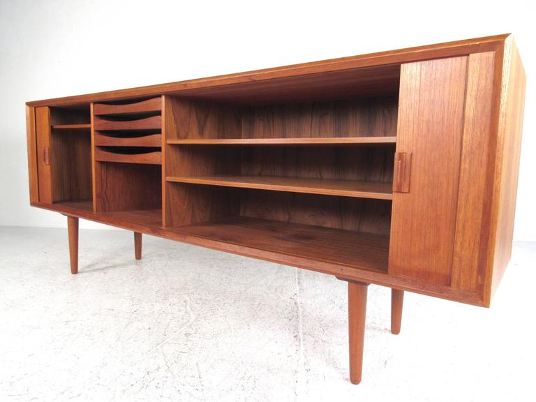 This stunning Danish Modern tambour sideboard features a beautiful teak finish, tapered legs and spacious interior cabinet space. Multiple shelves and felt lined drawers make this a versatile storage piece for any Mid-Century setting. Made in