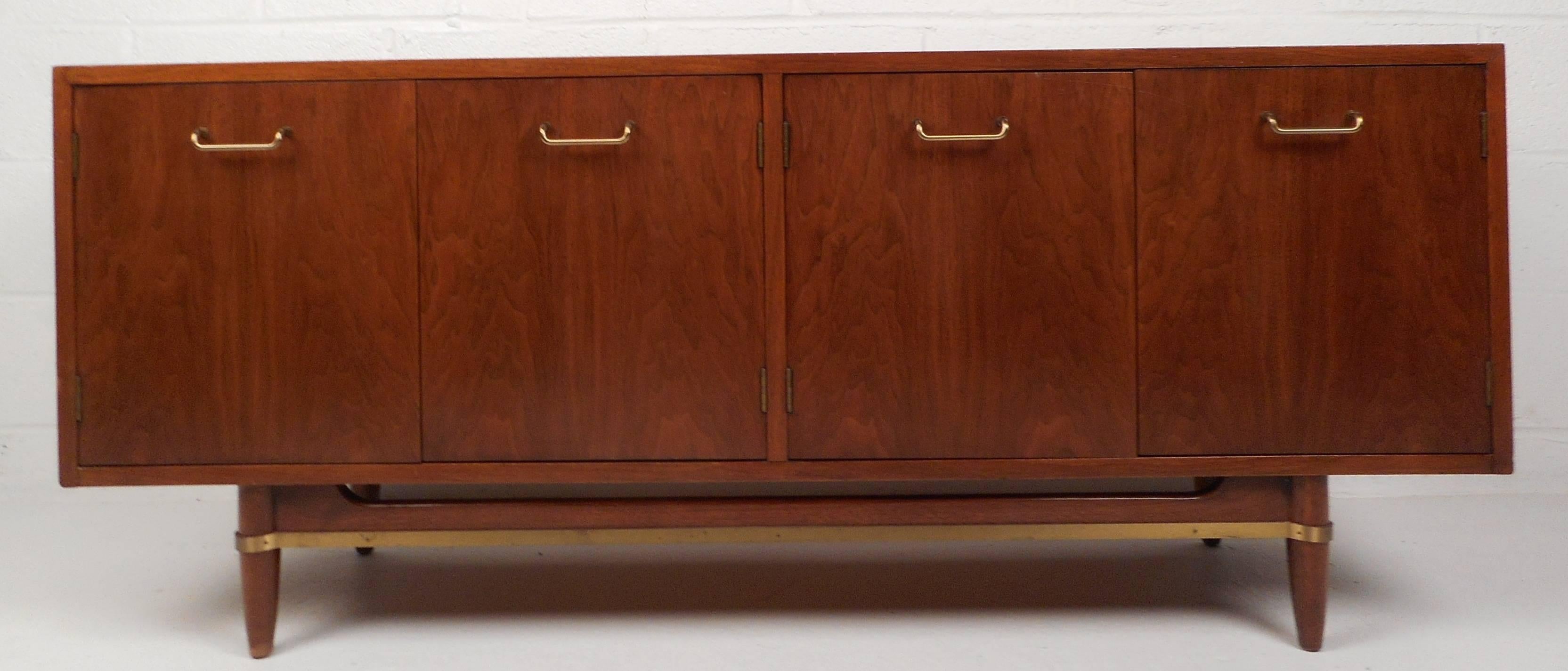 Stunning vintage modern small credenza features unique brass pulls and a vintage walnut finish. The stylish sculpted base has a brass trim stretcher and tapered legs. Two hefty storage cabinets and one drawer offer plenty of storage space without
