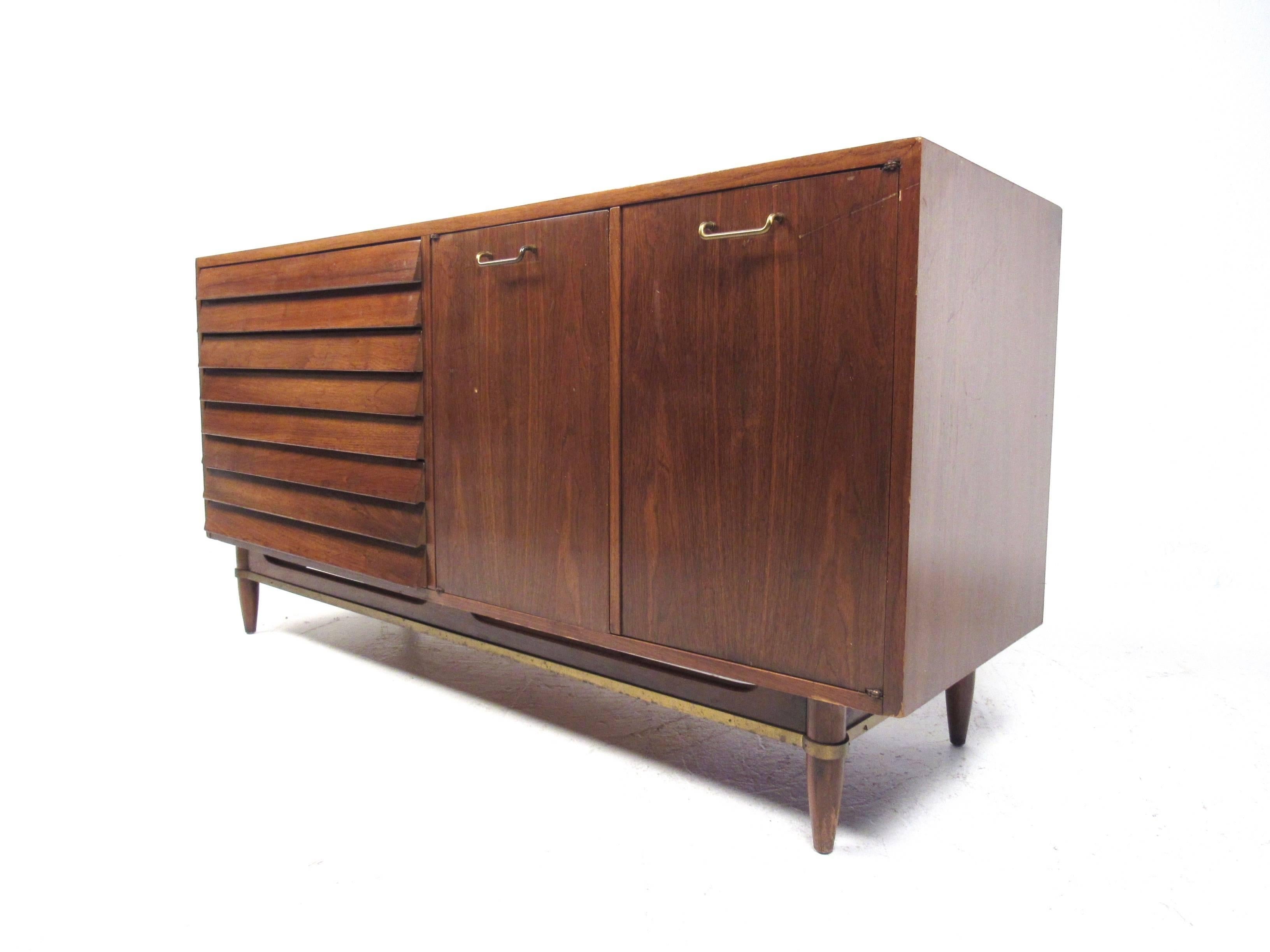 This beautiful vintage credenza features a unique walnut finish with louvered front drawers. Brass trim adds to the Mid-Century appeal of this miniature American of Martinsville storage piece, perfect size for smaller apartments with plenty of